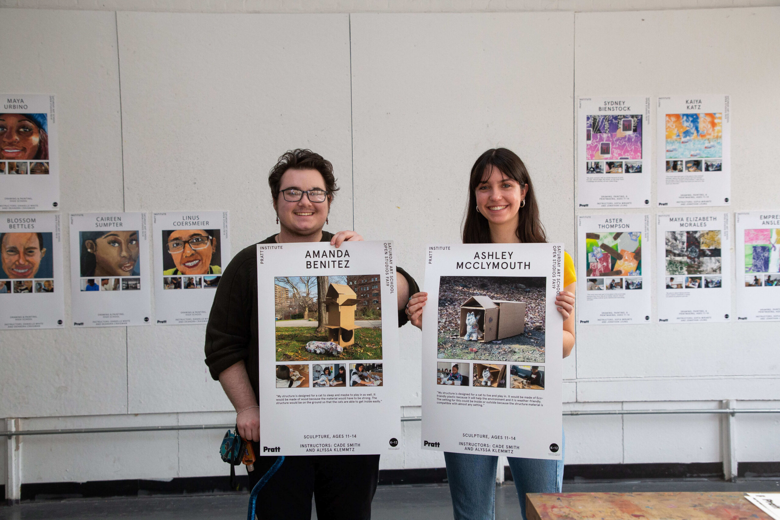 Two students present their work. They are standing with large posters at the front of a classroom. The posters contain images and text.