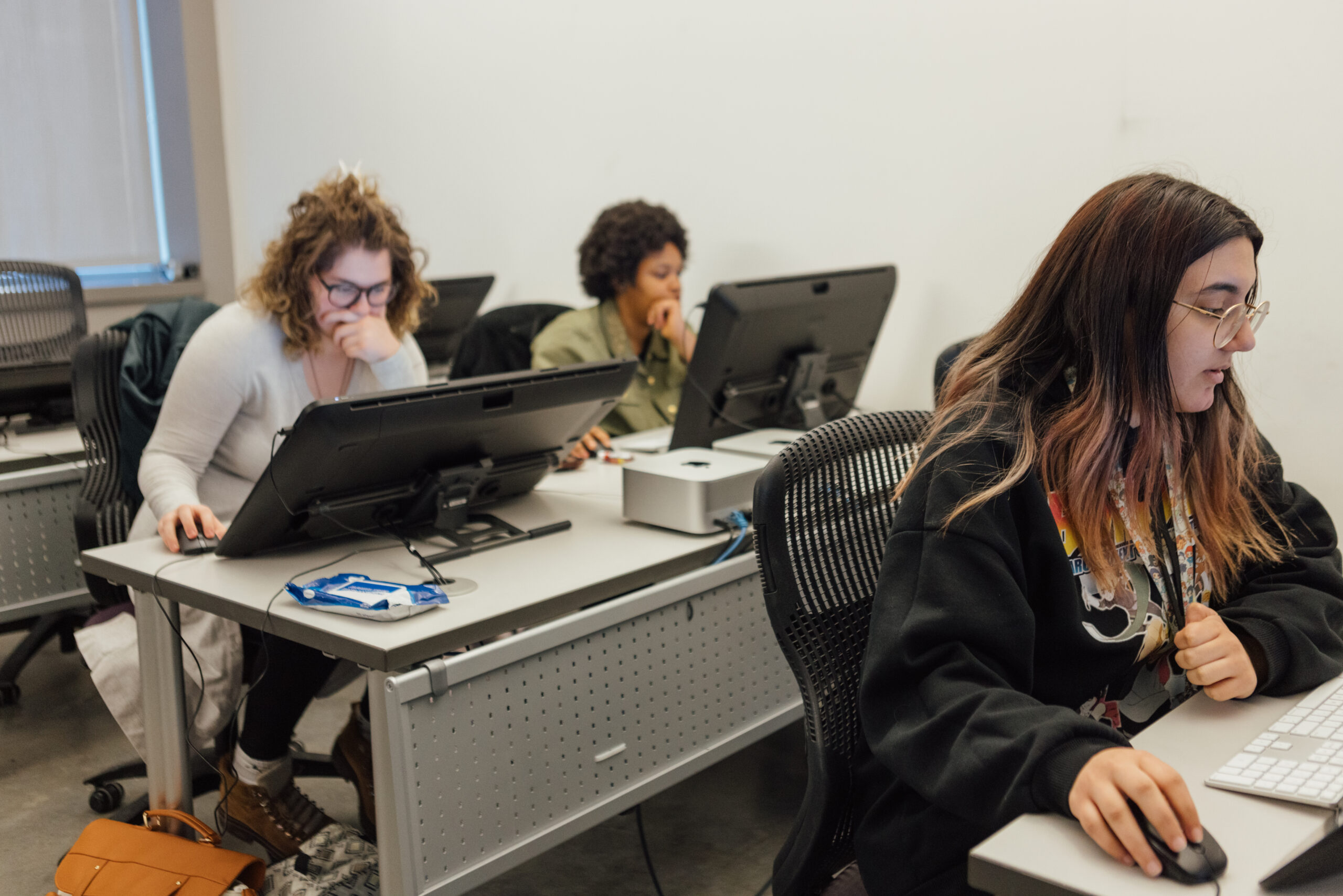 Three students are seated at desks in a classroom, each working intently on their own digital drawing tablets connected to desktop computers. The student in the foreground, with long, dark hair and glasses, is using a stylus to work on a tablet. The student in the middle has curly hair and is resting her hand on her chin, focusing on her screen. The student in the background has short curly hair and is similarly engaged with her work. The classroom is equipped with modern computer workstations and has a studious atmosphere.