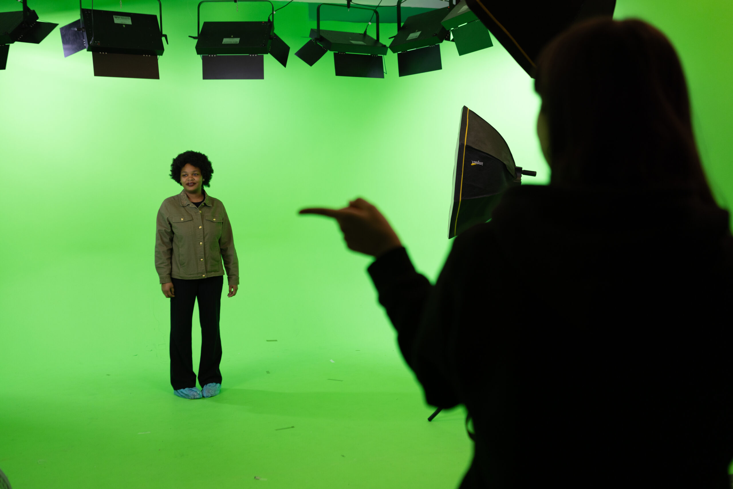 A person stands in a green screen room, facing the camera with studio lights hanging from the ceiling. They are wearing a green jacket and black pants with blue shoe covers. Another person, partially visible and out of focus in the foreground, is pointing towards the person on the green screen, seemingly giving directions or instructions. The scene suggests a studio environment where video or photo production is taking place.