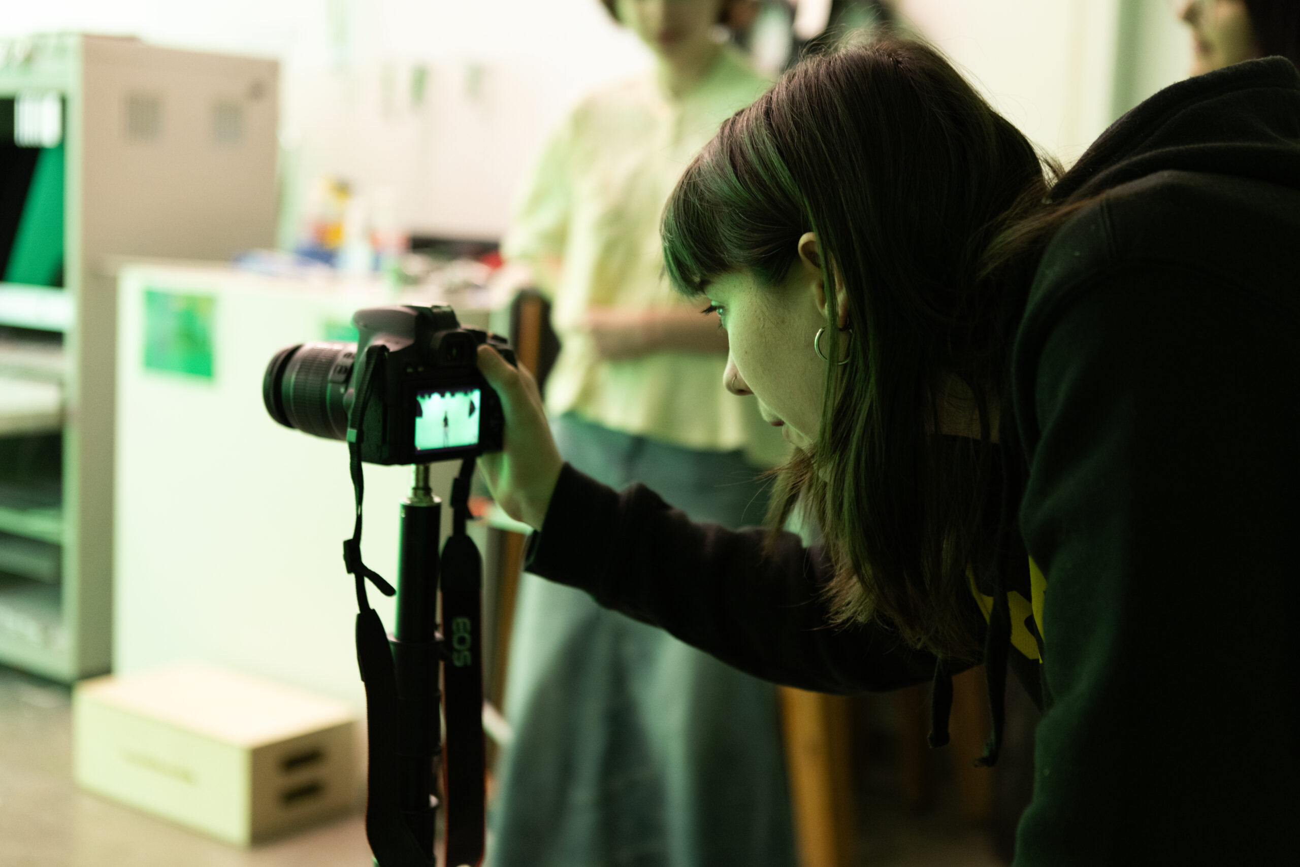 A person with long hair is looking through the viewfinder of a digital camera mounted on a tripod. They are focusing on the screen, which shows an image being captured. The scene is lit with a greenish hue, suggesting indoor lighting. In the background, there are other individuals and various equipment, slightly out of focus. The environment appears to be a studio or classroom setting.