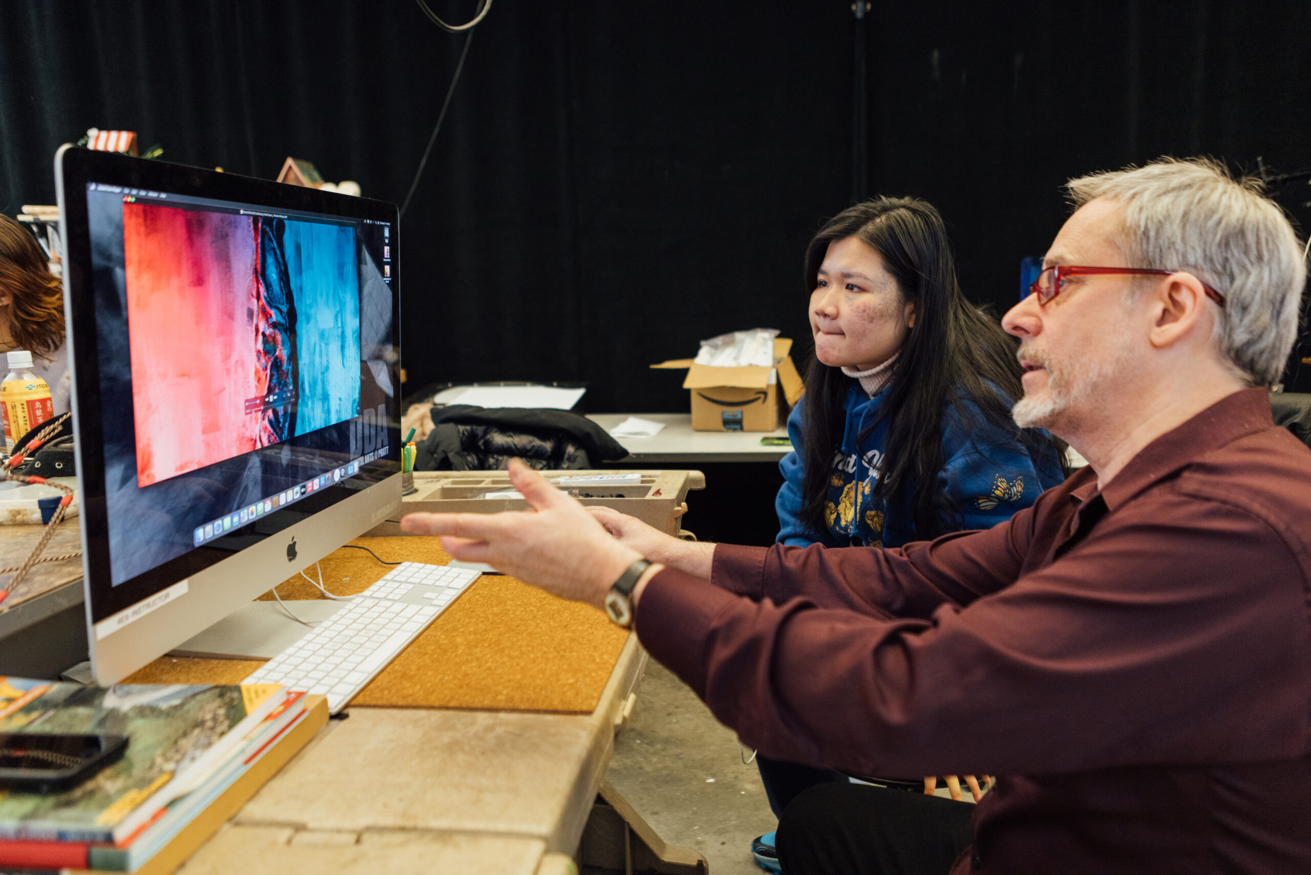 A man with gray hair and red glasses is sitting next to a young woman with long dark hair. They are looking at a large computer monitor displaying a colorful abstract image with red and blue hues. The man is gesturing with his hands while explaining something. The young woman is attentively listening. The background includes a black curtain, a cluttered table, and various objects. The setting appears to be a creative workspace or classroom.
