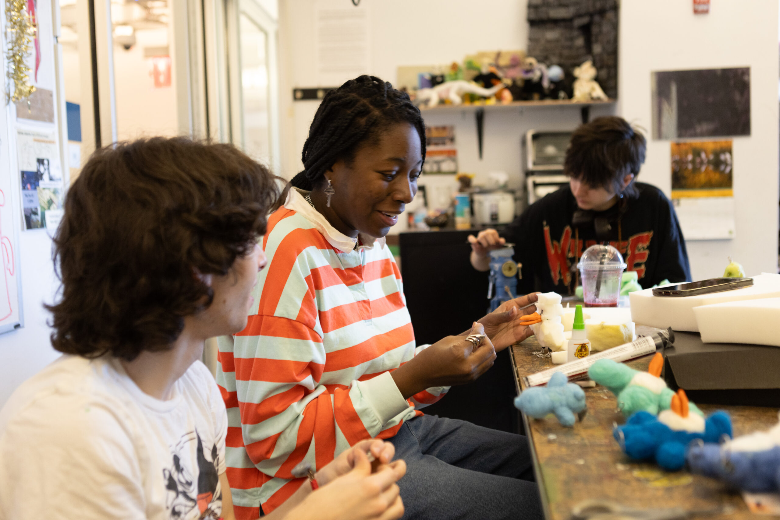 Three students are working on a project in a creative workspace. The student on the left, with curly hair, is listening to the middle student who has braided hair and is wearing a red and white striped shirt. The middle student is smiling while holding a small craft object. The third student, in the background, has short hair and is focused on their work, with various craft supplies and a drink on the table. The room is decorated with art materials and projects.