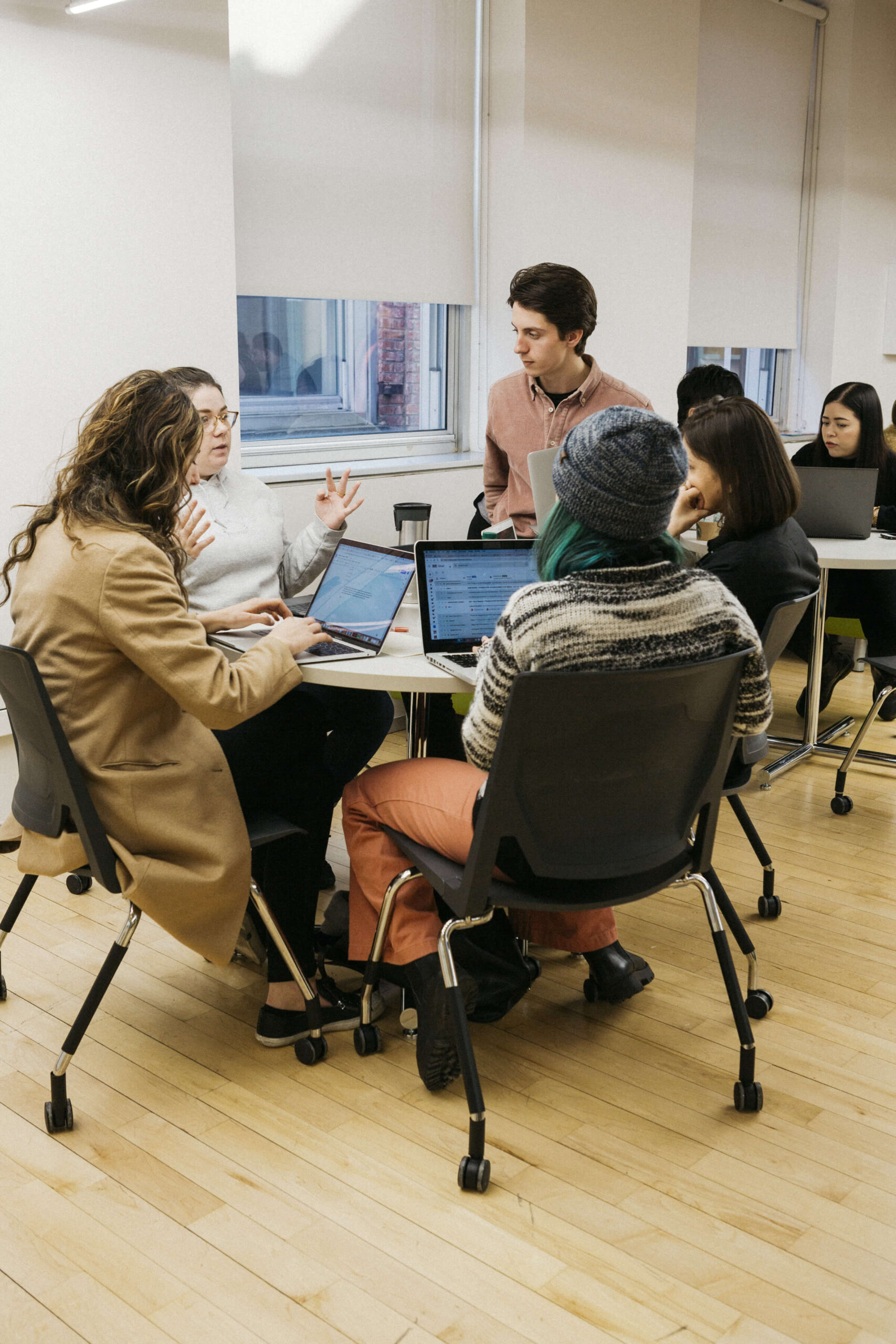 A group of student sit around a round table as they discuss. They all have laptops on the table. They are sitting on chairs with wheels.