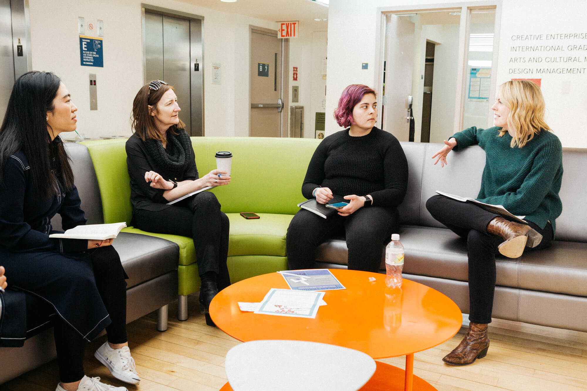 A group of students sit on a multicolored couch as they discuss. There is an orange coffee table in front of them with documents resting on top.