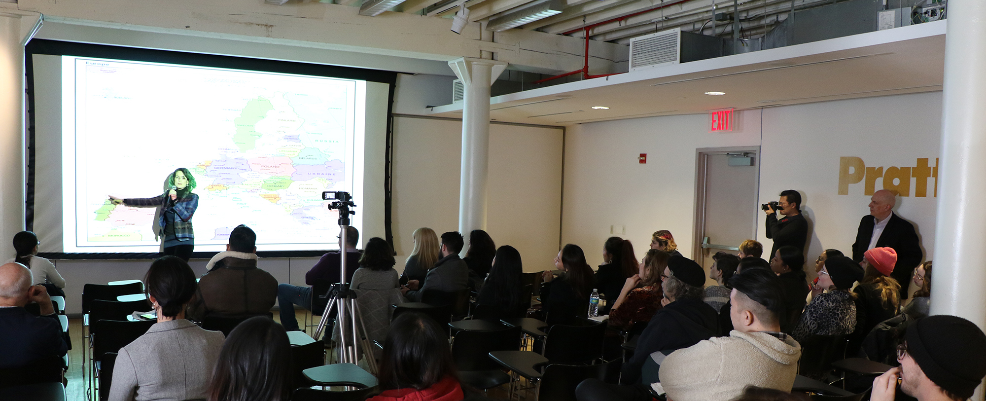 A woman presents a slide with a map to a large crowd inside a meeting hall at Pratt.