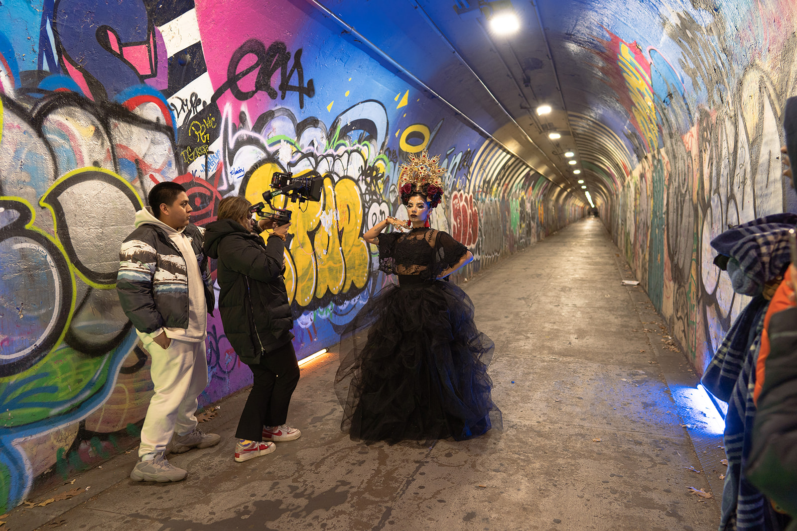 woman in long hallway, wall covered in graffiti, with man holding camera pointed at her