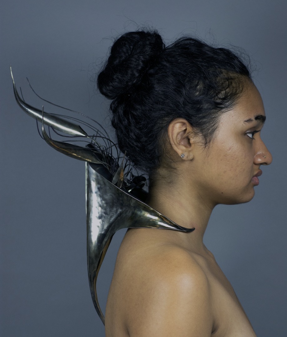 A person in side profile wears a silver-toned, abstracted form from the back of their neck, from which metal strands and feather-like forms protrude outward.