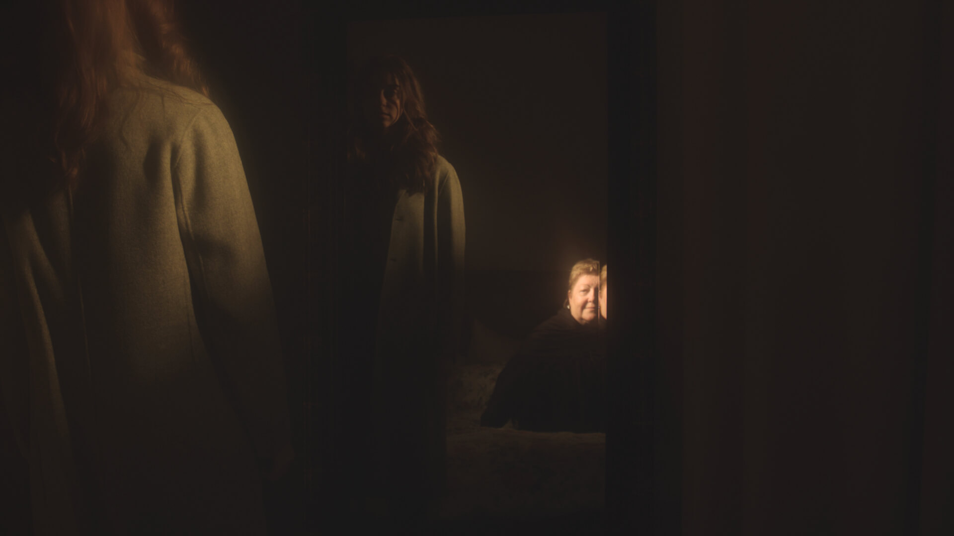 Movie still of a woman looking at her reflection in a dark room. She is wearing a white robe.