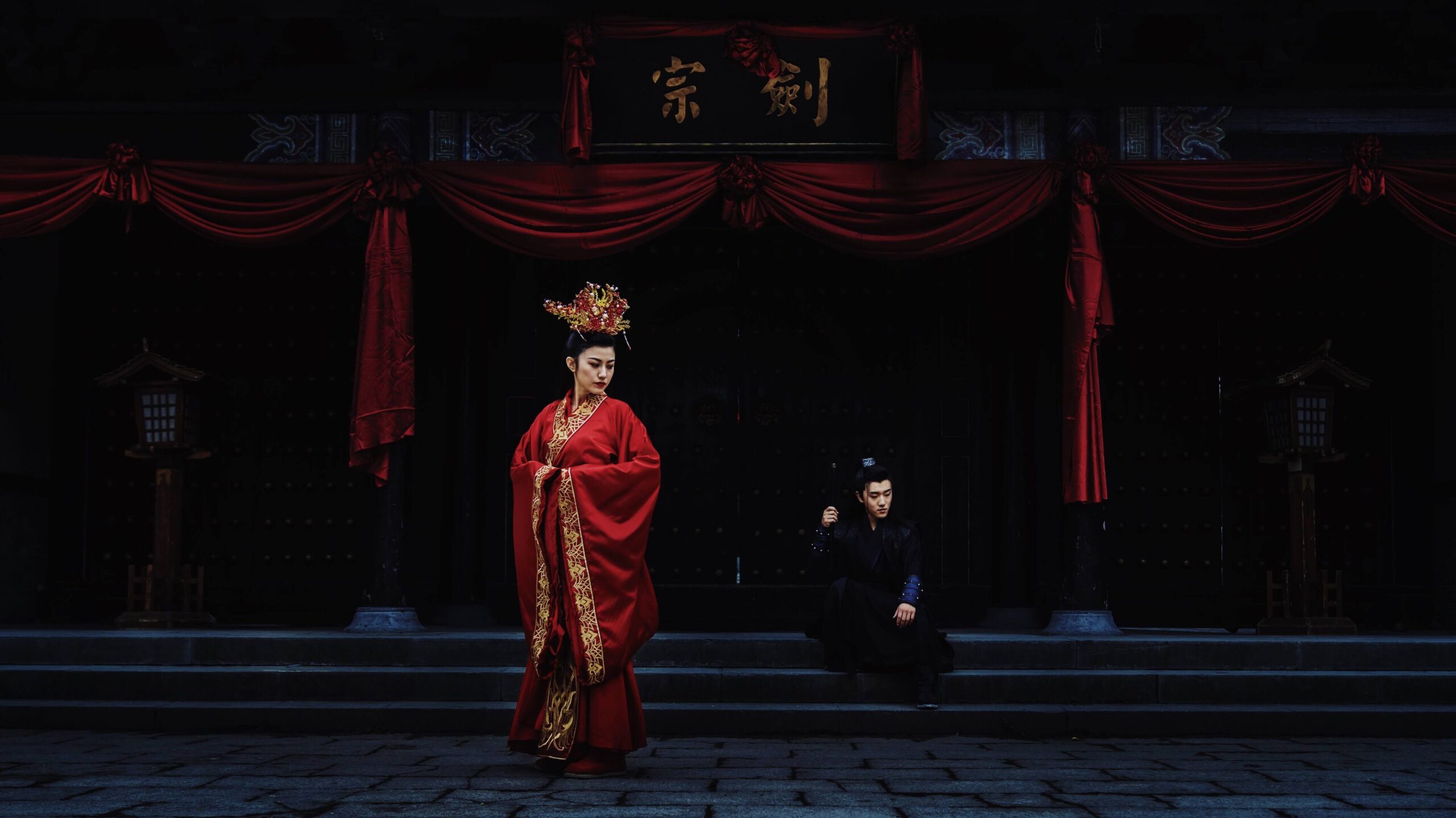 A woman in a red traditional Chinese dress stands in front of stairs in a courtyard. There is a man sitting on the steps wearing a black traditional Chinese tunic.