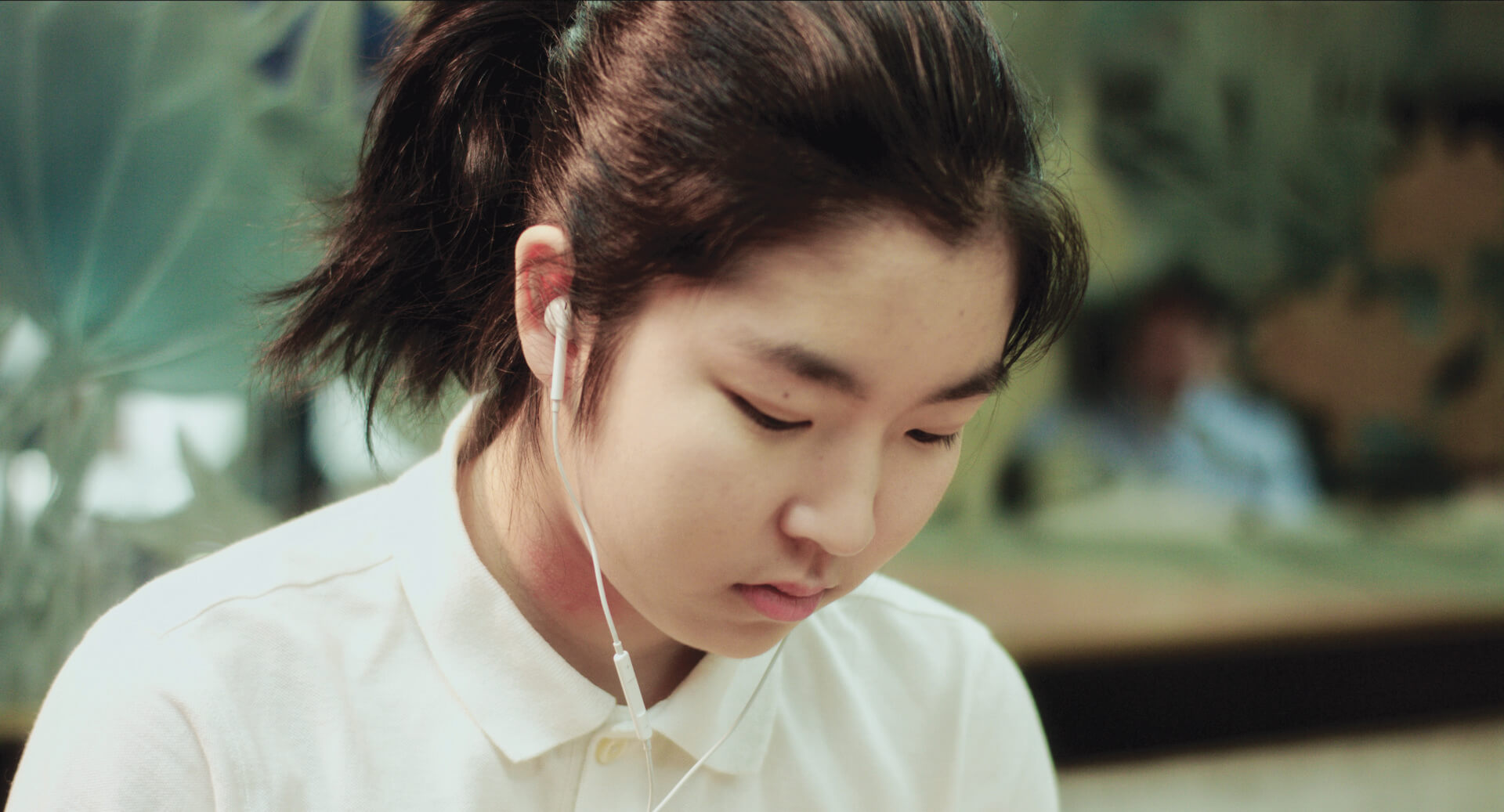 A girl's face is in focus as she listens through her earphones and looks down with an emotionless face.