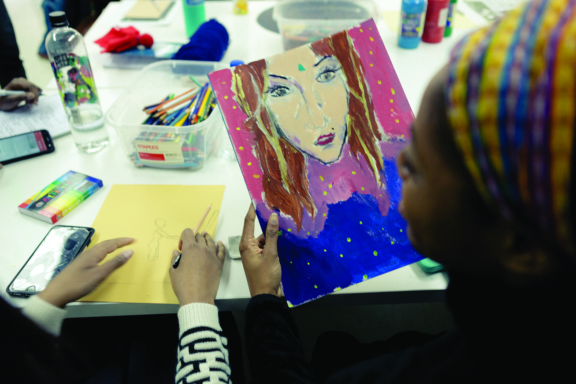 student holding a hand-drawn painting, showing it to another person, in studio room, brushes, painting supplies in the background