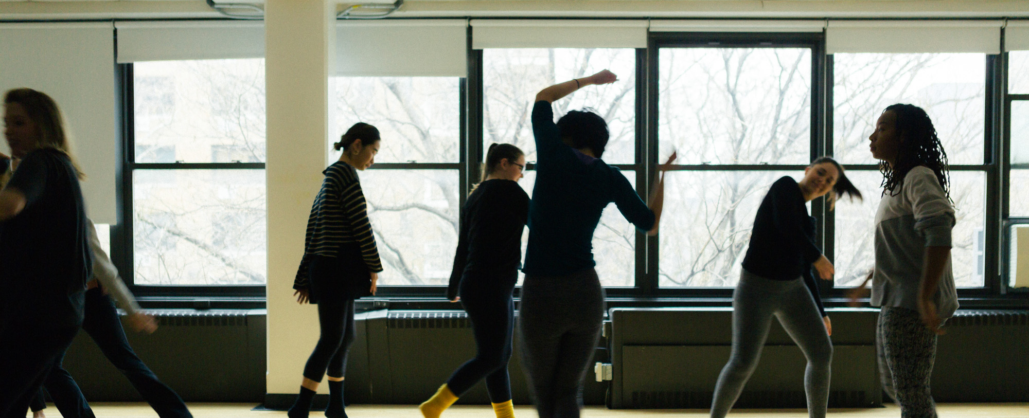 A group of dancers practice in a room with windows in the background.
