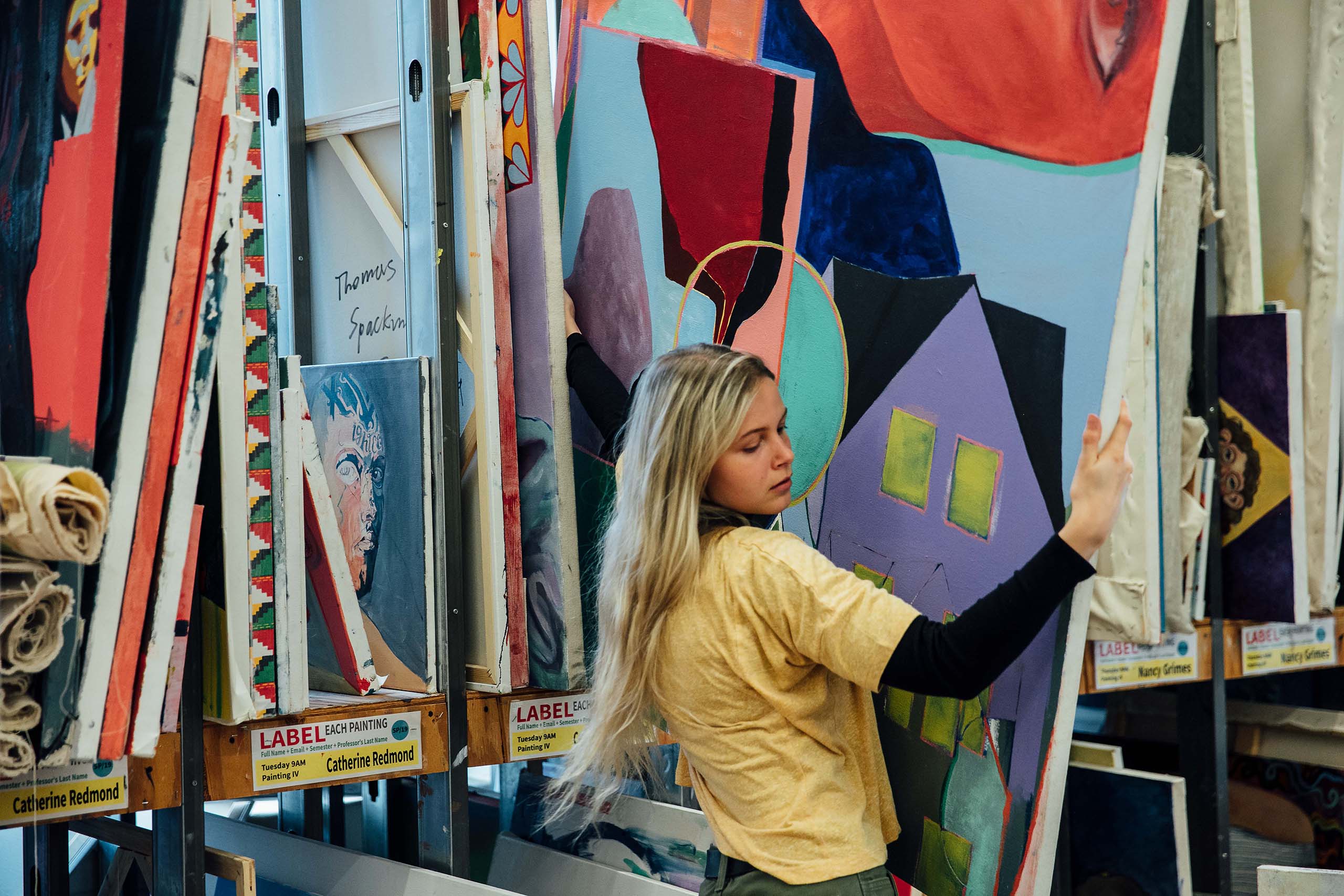 A student carries a canvas in a studio filled with various multicolored paintings.
