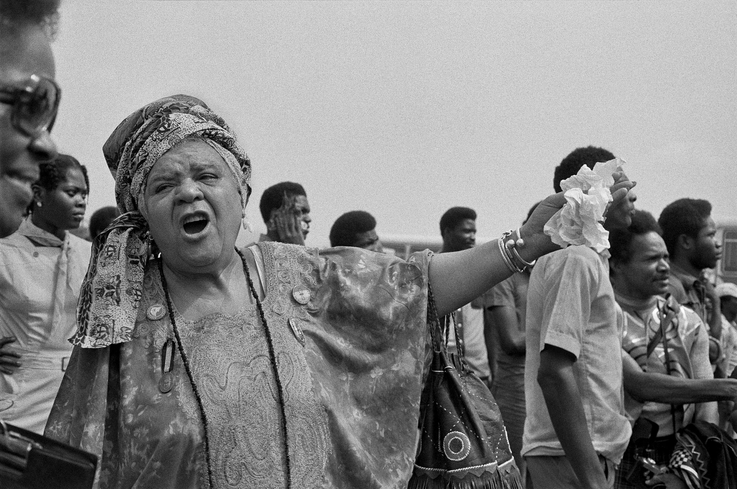 Queen Mother Audley A. Moore, in a knotted headdress and patterned gown adorned with badges, speaks to a companion, gesturing with her left arm