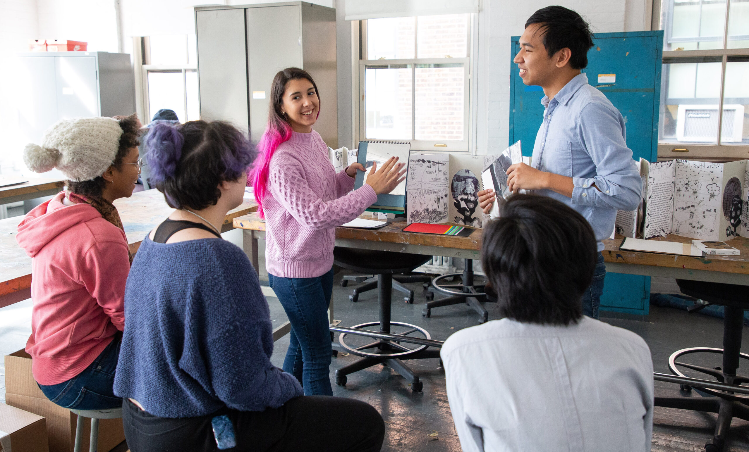 Two people are standing presenting to three people sitting. They are showing works of art that are propped up on a work table.