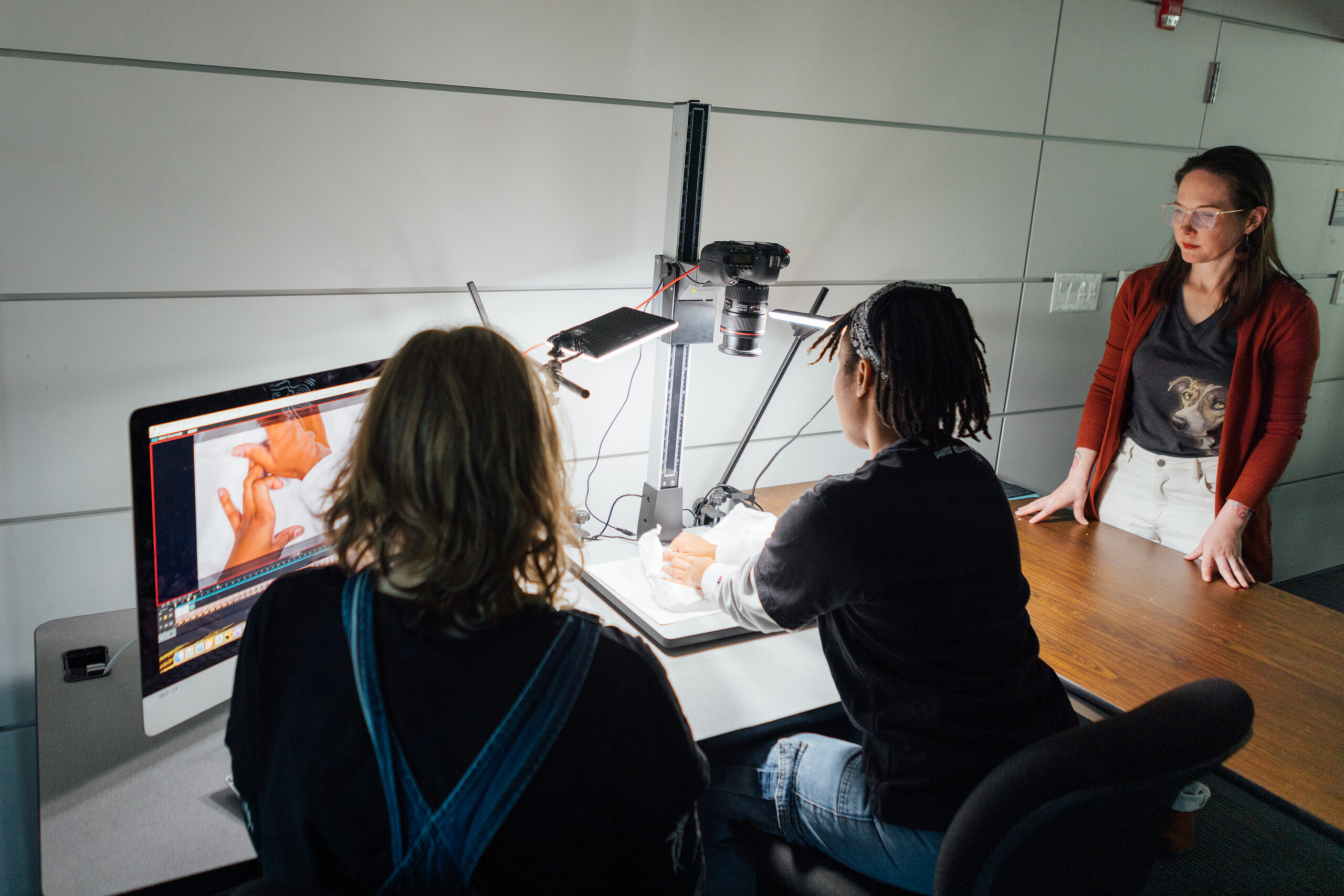 In a dimly lit room, two people are seated at a table working on an animation project. They are using a camera setup with adjustable lights to capture images of hands manipulating objects on a white surface. One person, with shoulder-length hair, is looking at a computer monitor that displays the captured images. The other person, with short hair and a headband, is focused on the setup. A third person, standing at the end of the table, is observing their work, wearing glasses, a dark T-shirt with a dog print, and a red cardigan. The scene conveys a collaborative environment focused on stop-motion animation or a similar project.