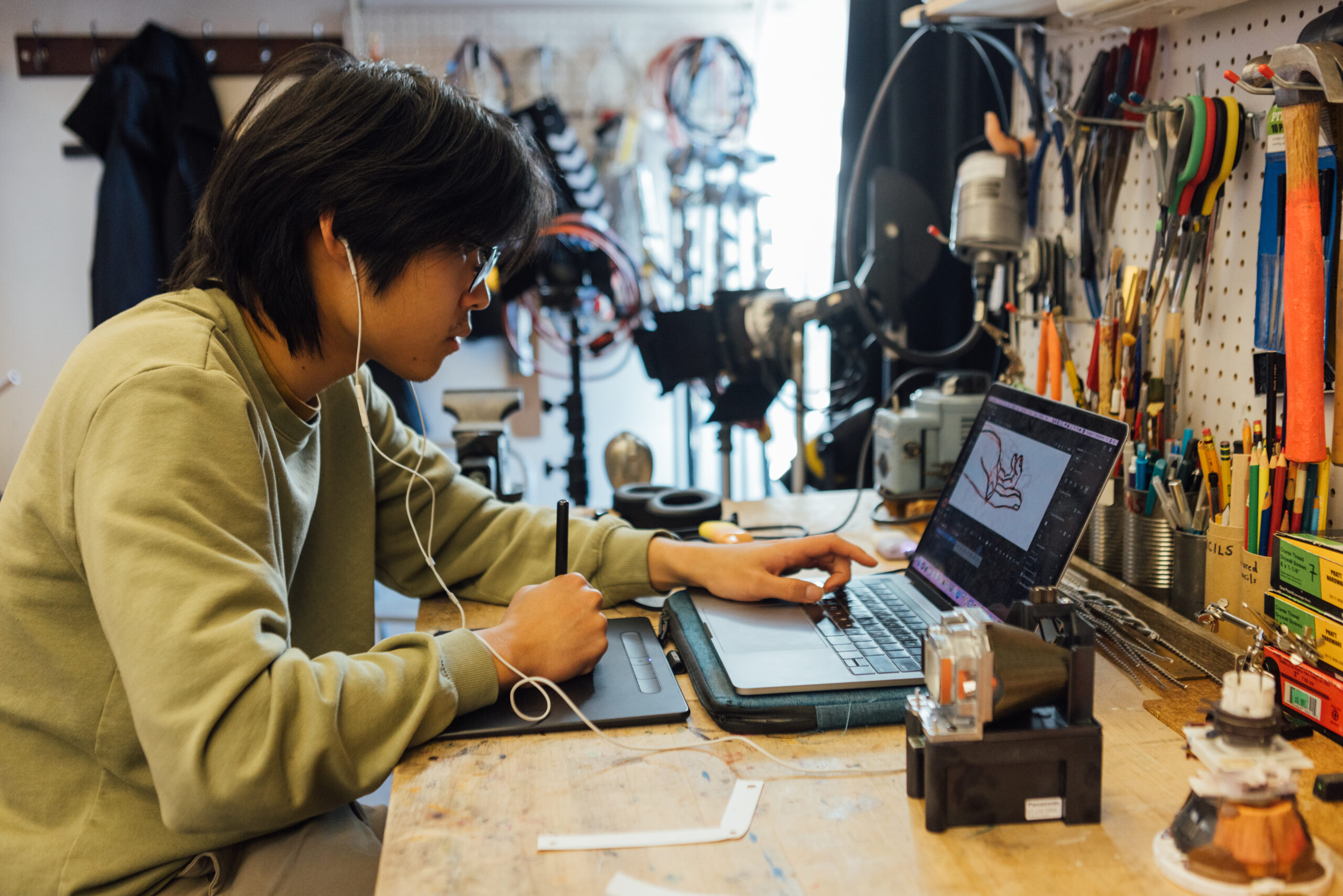 A person with shoulder-length hair, wearing glasses and a light green sweatshirt, is sitting at a cluttered workbench in a workshop. They are focused on a laptop while drawing on a graphics tablet, with the screen displaying an animation project. The workbench is filled with various tools, including pliers, screwdrivers, and wrenches, organized on a pegboard in the background. The scene suggests a creative and technical environment, emphasizing the process of digital animation and design.