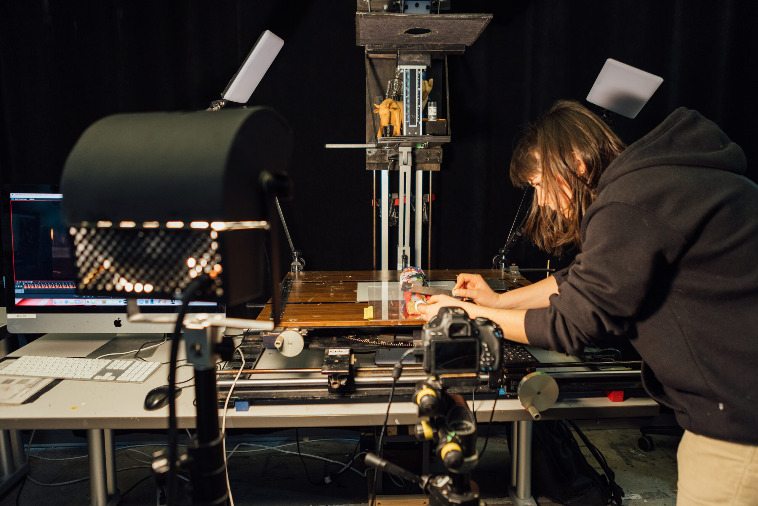 A person with shoulder-length hair, wearing a black hoodie, is working on a stop-motion animation setup. They are carefully adjusting a small, colorful puppet on a wooden table, surrounded by professional lighting equipment and a mounted camera. A computer with an animation software interface is visible on the desk in the background, alongside various tools and equipment used in the animation process. The scene depicts a detailed and technical environment, highlighting the meticulous work involved in creating stop-motion animation.