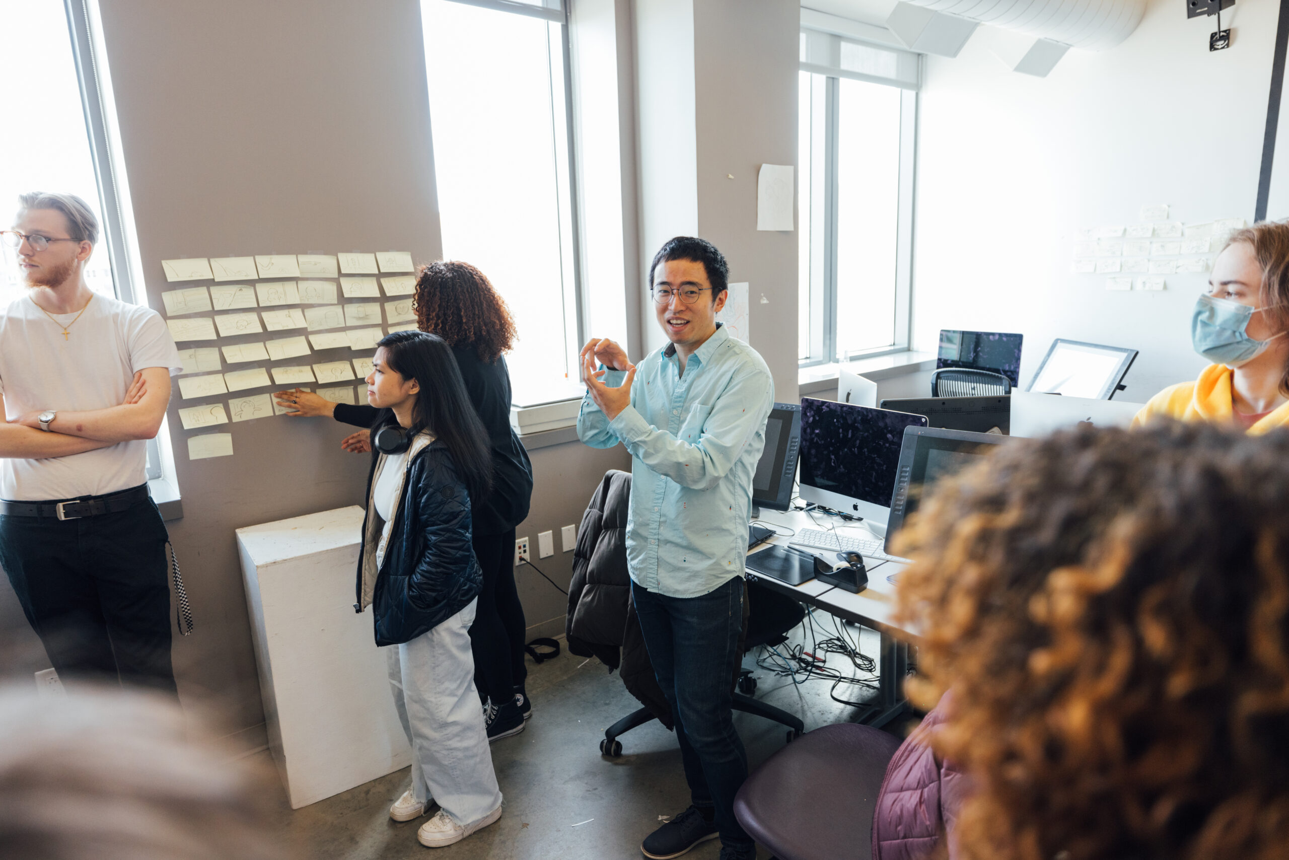 A group of people are engaged in a collaborative discussion in a modern office setting. One person, wearing a light blue shirt and glasses, is gesturing with their hands while speaking. To their left, a woman with long black hair and wearing a black jacket is observing sticky notes on a wall. Another person, with curly hair, is adding or adjusting sticky notes on the same wall. On the left side of the image, a man in a white shirt with a cross necklace stands with his arms crossed, looking towards the discussion. In the background, there are large windows providing natural light, several computer workstations, and another person with a face mask and a yellow hoodie, focusing on the discussion. The scene suggests an interactive and collaborative environment, possibly a brainstorming session or team meeting.