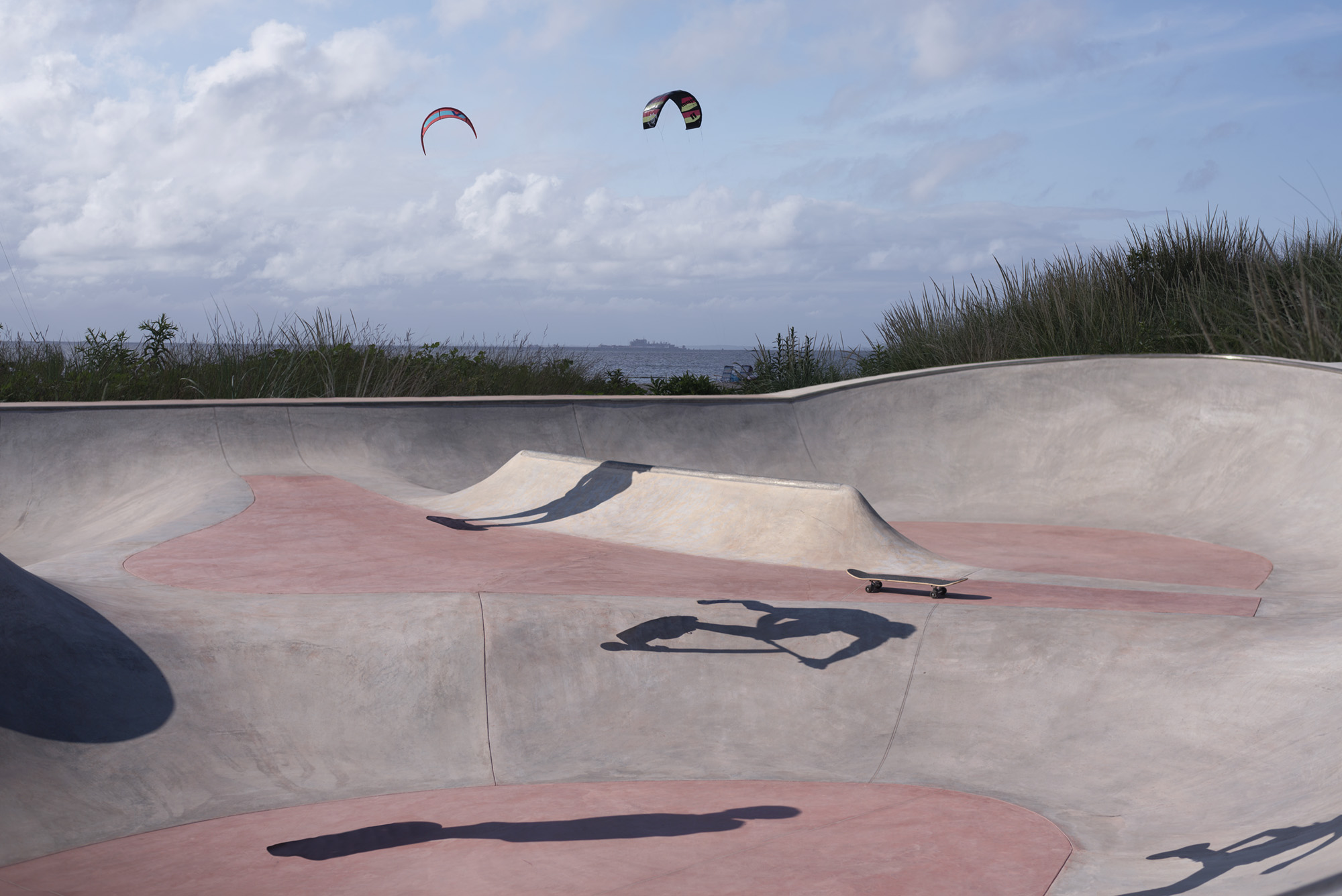 An empty skatepark the color of coral. The shadows of people are present on the ground though the figures that belong to them cannot be seen as though they have been removed from the photograph. There are two paragliders in the air.