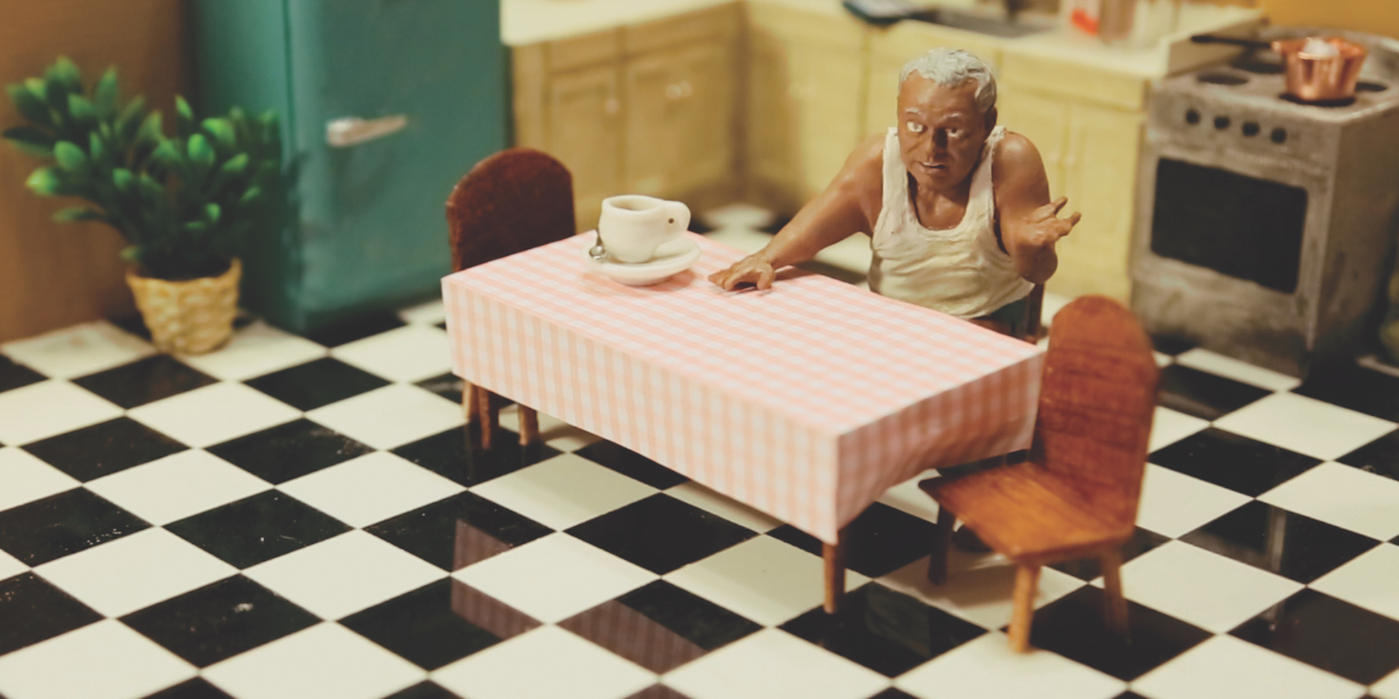 Figurine of man in white tank shirt sitting in a model of a kitchen with checkerboard floor