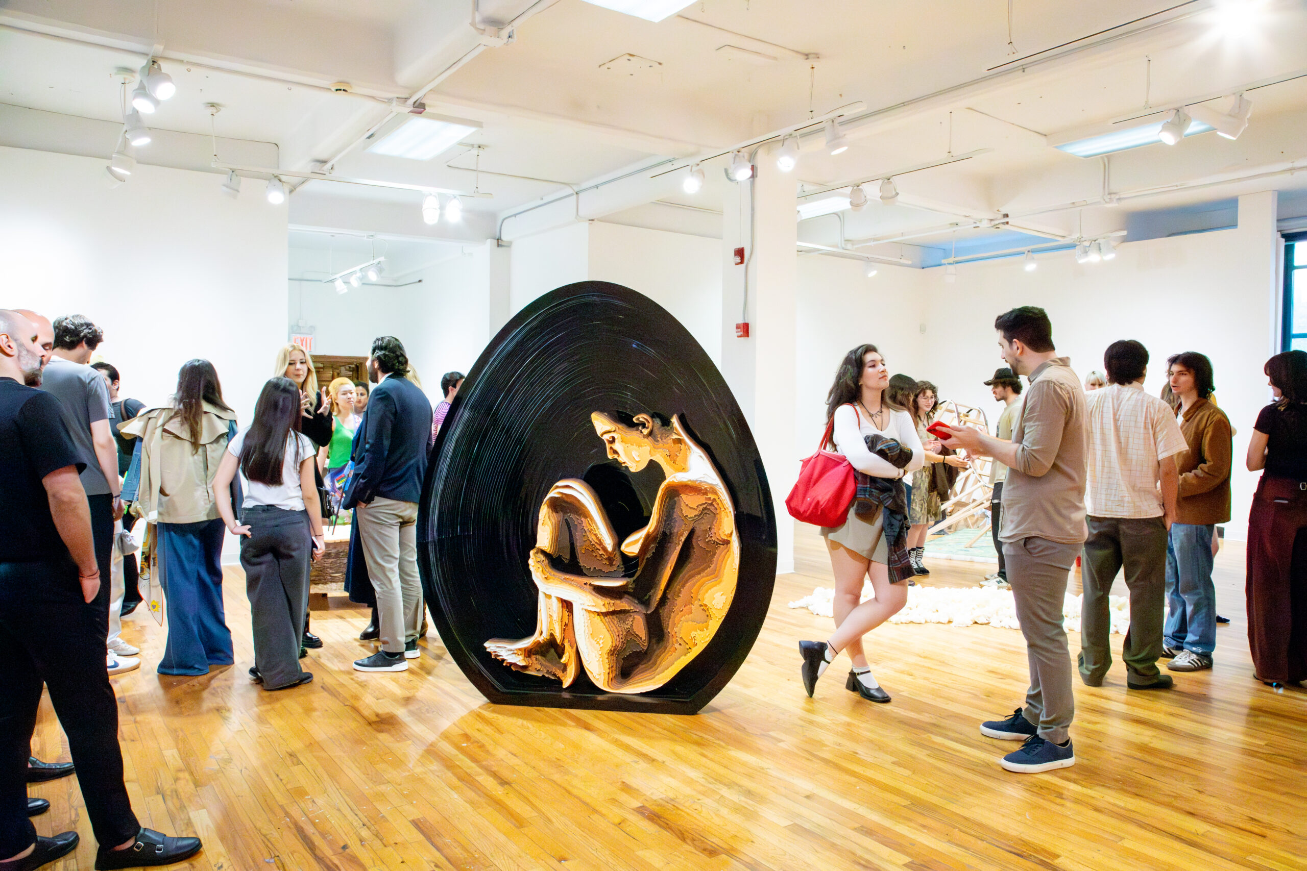 Gallery opening with visitors standing and talking around a large sculptural installation shaped like a dark egg, featuring a seated human figure inside, in a bright room with wooden floors.