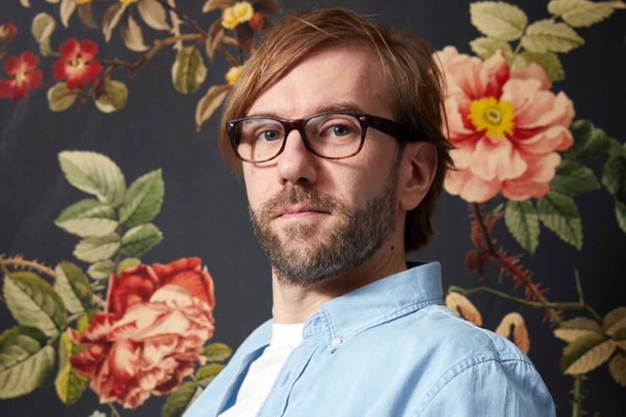 headshot of Jeremie Perin, wearing glasses, with beard and dress shirt, standing against a backdrop with flowers,