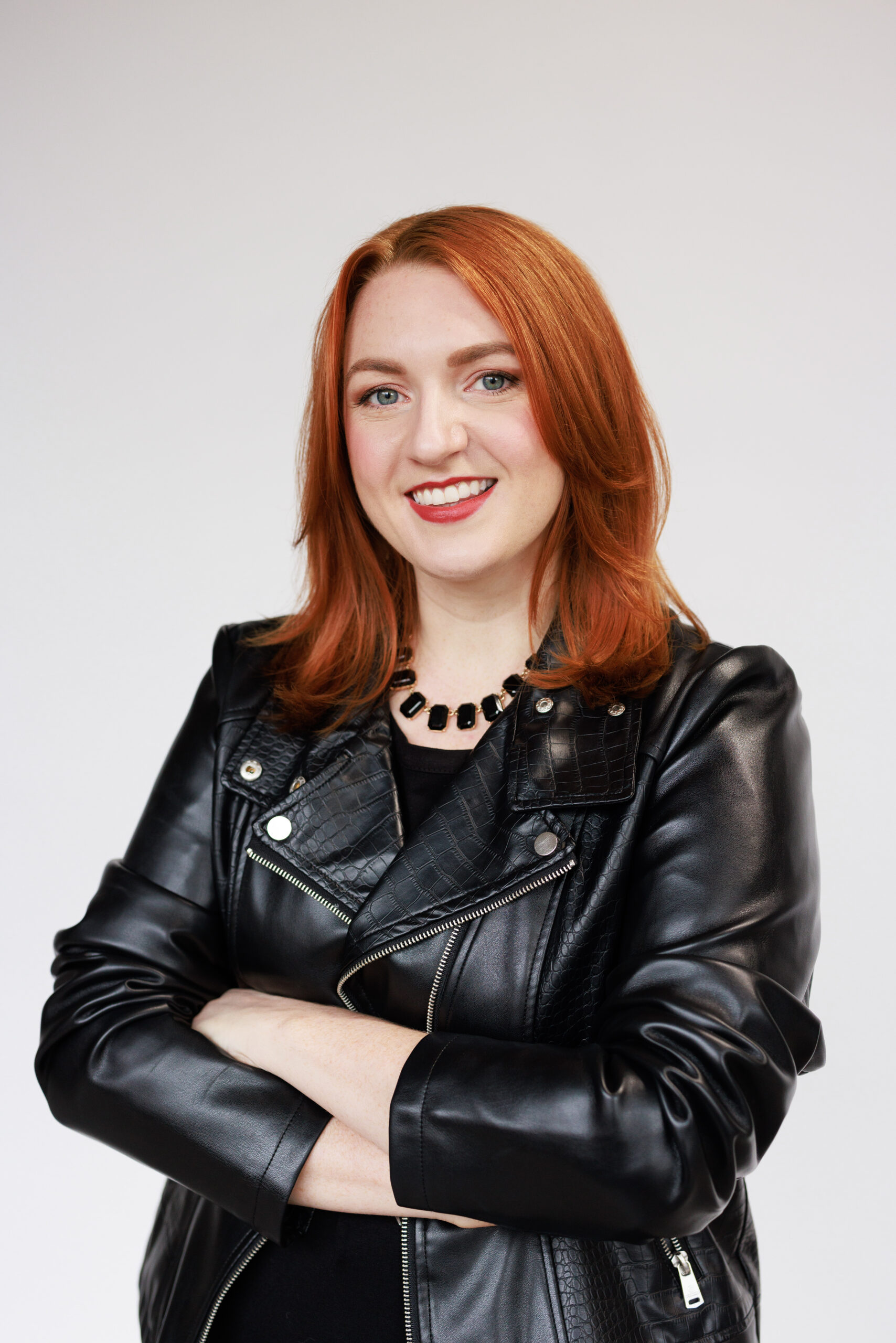 A headshot of a red head woman in a leather jacket in front of a white background.