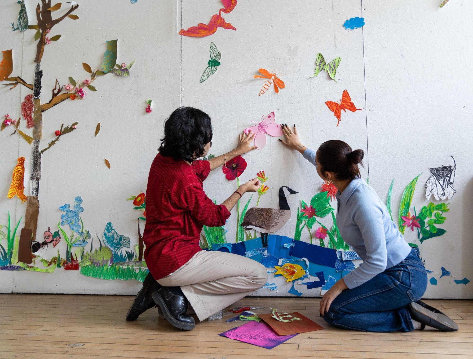 Two people kneeling on the floor work on creating a large colorful mural featuring birds, trees, and flowers. One person in a red shirt adjusts a paper butterfly on the mural while the other, in a blue shirt, reaches up to assist.