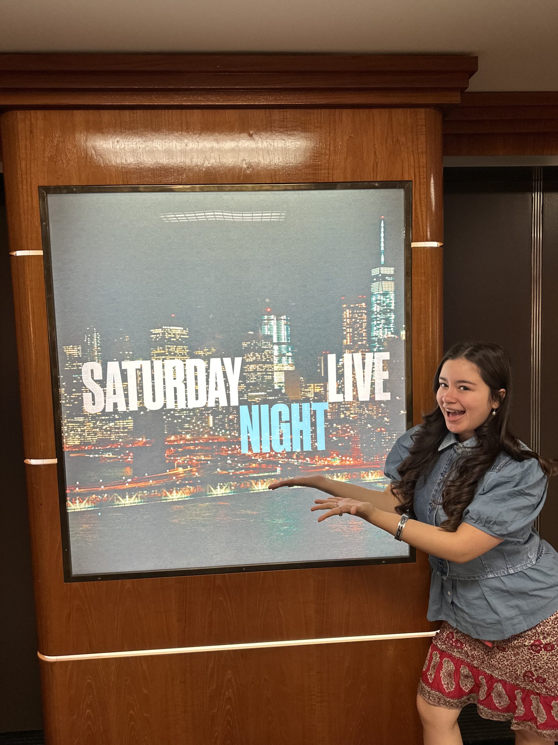 A woman smiles and holds her hands out in front of a sign. that says Saturday Night Live.