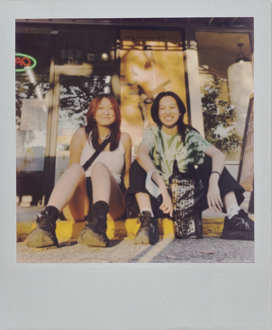Polaroid of Eileen and Grace, smiling as they sit on a curb in front of a store under dappled light