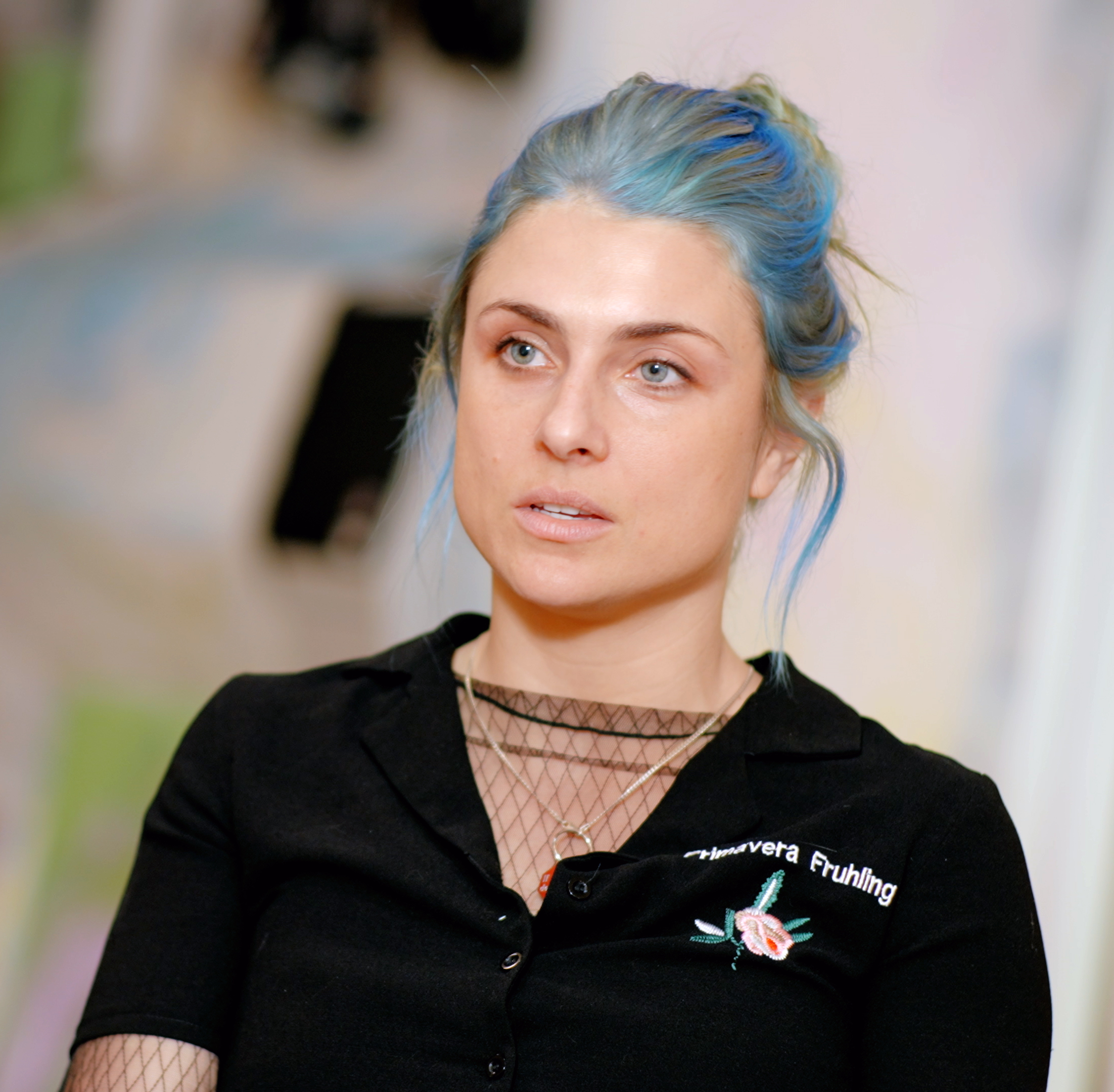 Portrait of a person with blue hair wearing a black shirt with floral embroidery and a mesh undershirt, against a blurred indoor background.
