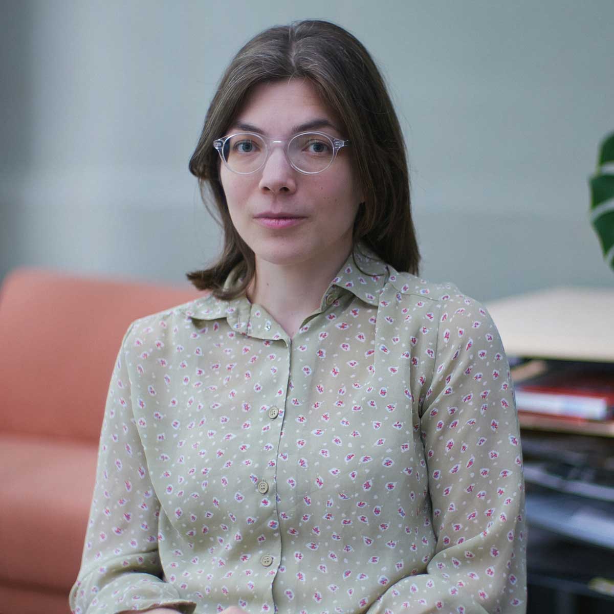 A woman with shoulder-length brown hair and clear-framed glasses sits indoors. She is wearing a light green button-up shirt with a small floral pattern. Behind her is an orange couch and a table with stacked papers and a plant partially visible. The setting appears to be an office or lounge area.