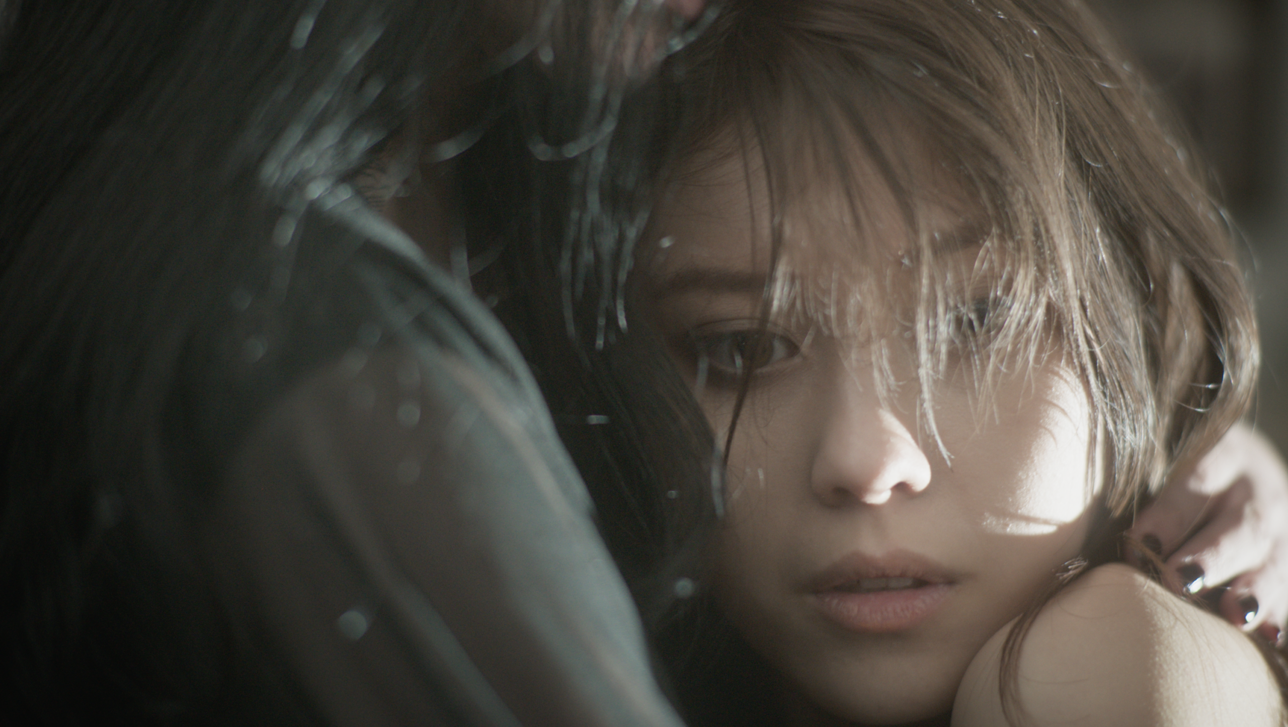A close-up of a young girl with long, dark hair, looking directly at the camera with a contemplative expression. Her hair has some strands falling over her face. In the background, a figure is partially visible, with dark hair and an arm around the woman's shoulder, creating an intimate atmosphere. Soft lighting enhances the emotional tone of the image.