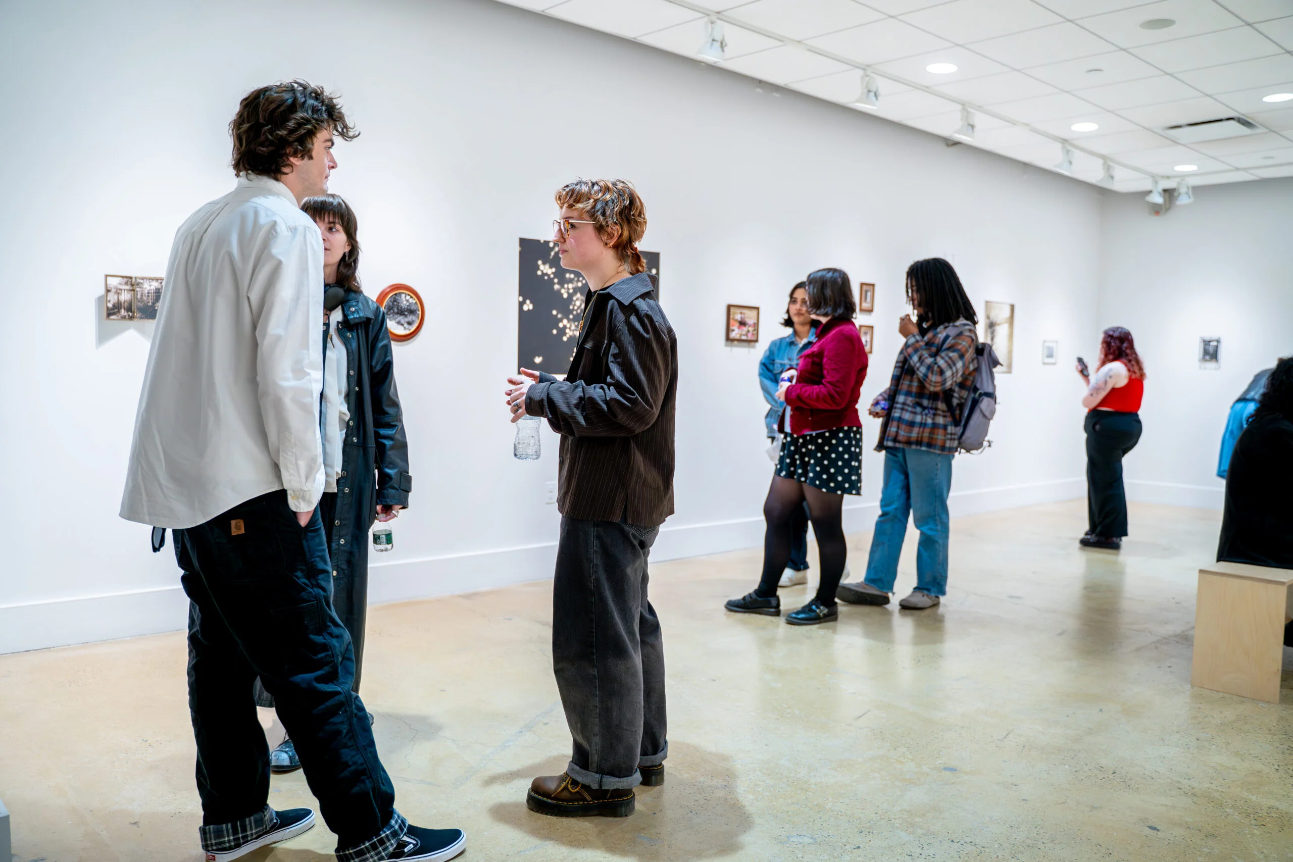 Visitors stand and converse inside a white-walled gallery during a photography exhibition. In the foreground, three people face one another in discussion, one holding a clear water bottle. Additional visitors are spaced along the gallery wall in the background, viewing small framed artworks and wall-mounted pieces. The room features polished concrete floors, overhead track lighting, and evenly spaced artworks arranged at eye level, creating an open, informal exhibition environment.