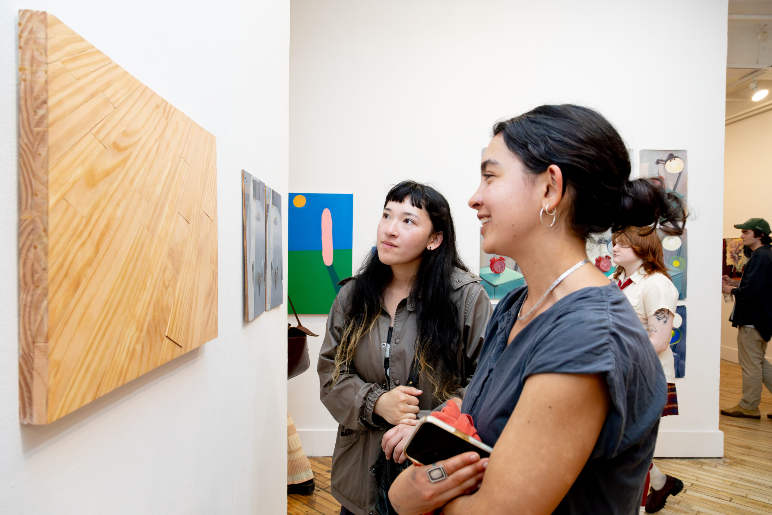 Two gallery visitors stand closely together viewing a wooden wall-mounted artwork with visible grain and geometric construction. One person looks attentively at the piece while the other smiles, holding a phone and folded fabric in their arms. Behind them, additional colorful paintings are displayed on white walls, and other visitors move through the exhibition space. The scene captures casual conversation and focused engagement with contemporary artworks in a well-lit gallery.