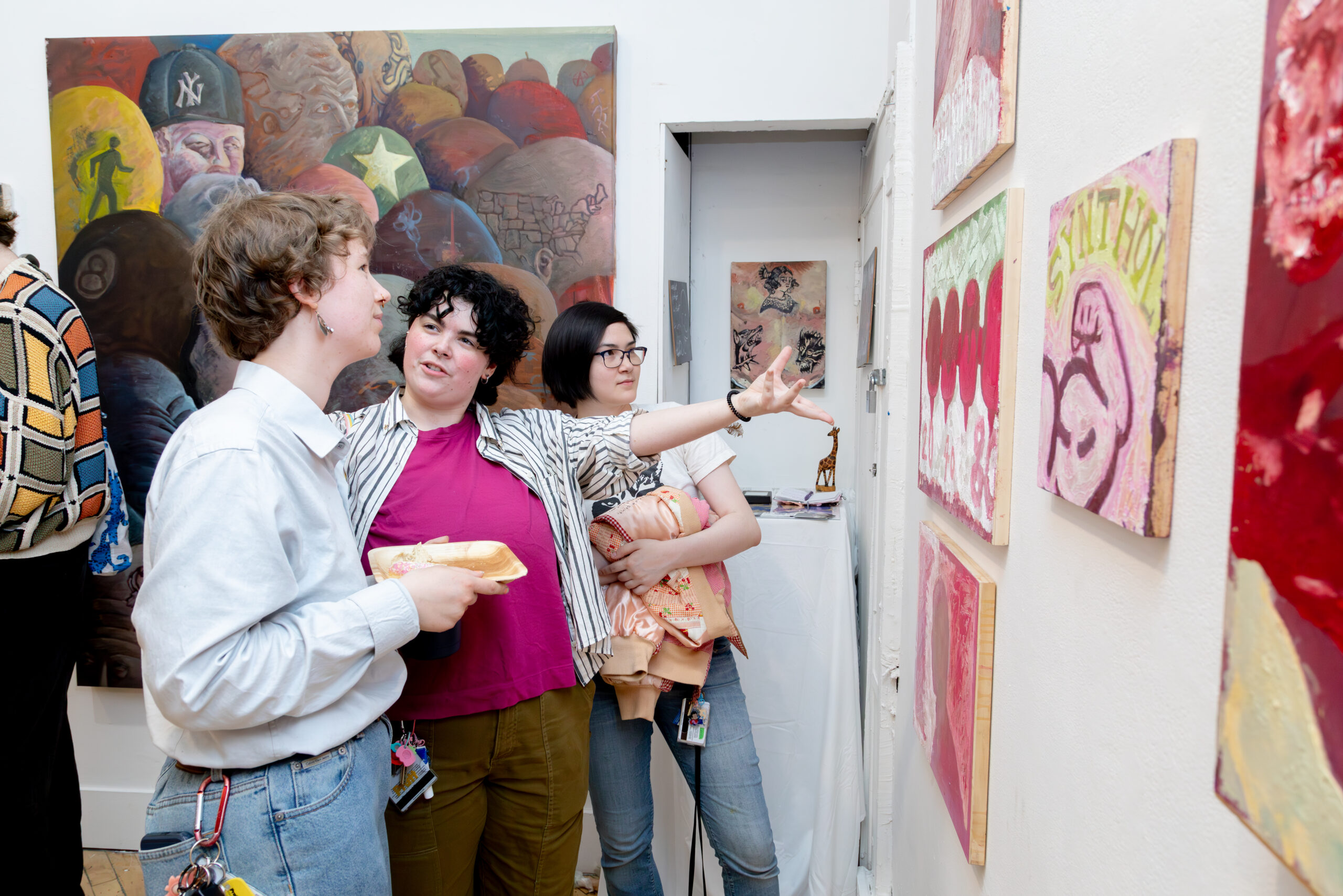 Three gallery visitors stand closely together in a painting exhibition, discussing artworks mounted on a white wall. One person gestures with an outstretched hand toward a series of small, colorful abstract paintings, while another holds a plate of food. A large figurative painting with layered imagery fills the background behind them, and additional artworks are visible through a doorway. The scene conveys active conversation and engagement with the exhibited paintings in a crowded gallery setting.