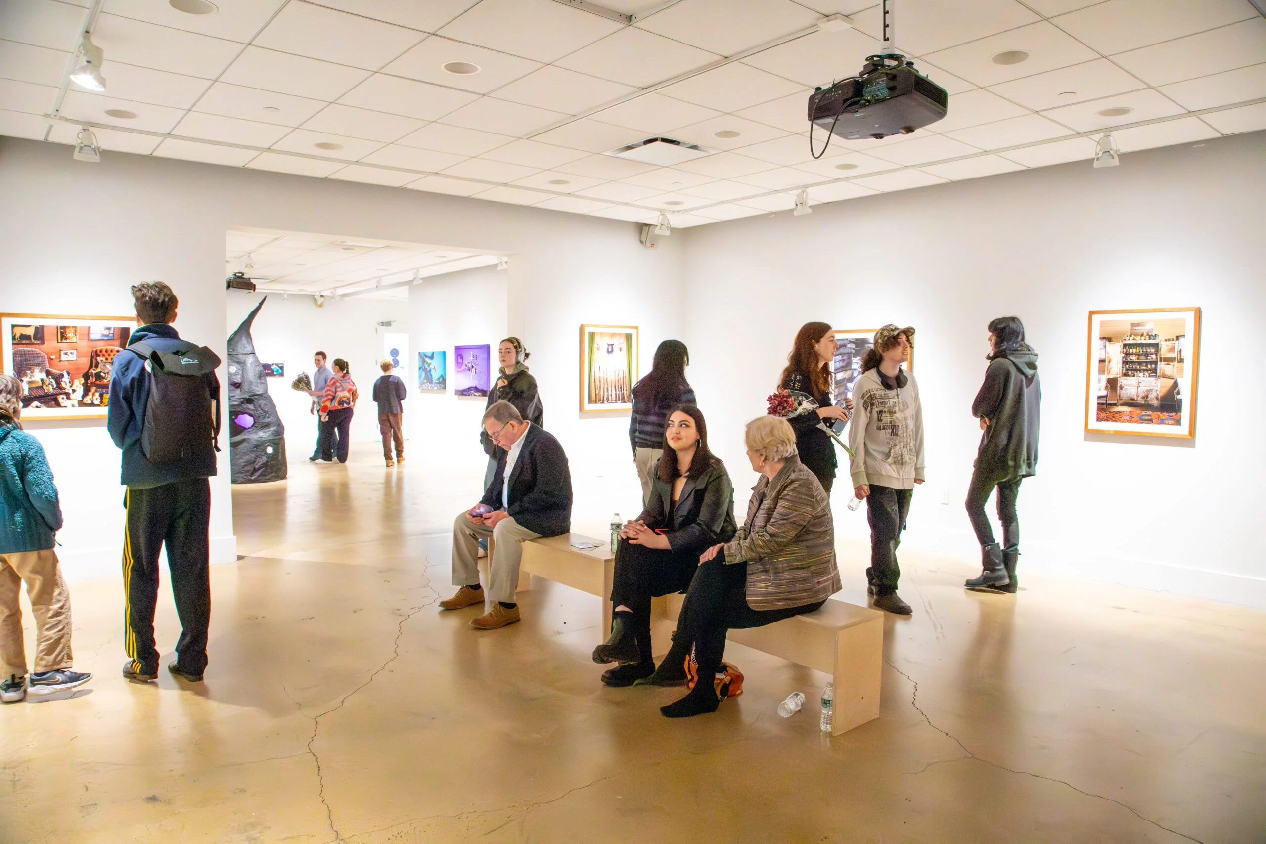 A wide view of a gallery exhibition space with multiple visitors dispersed throughout the room. Several people sit on a central wooden bench while others stand and converse or view framed photographic works mounted on white walls. A sculptural installation with an irregular vertical form is visible in the background near an open doorway connecting adjacent rooms. Overhead track lighting and a ceiling-mounted projector illuminate the space, emphasizing the active, social atmosphere of an art exhibition setting.