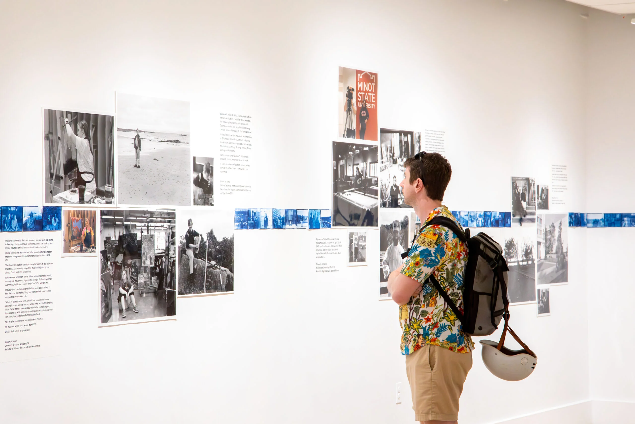 Gallery visitor wearing a patterned short-sleeve shirt, tan shorts, and a backpack stands in profile, closely examining a wall-mounted photographic installation. The exhibition consists of black-and-white documentary photographs, short blocks of wall text, and a continuous horizontal strip of small blue-toned images running across the wall at mid-height. The viewer holds a light-colored bicycle helmet at their side. The gallery space is brightly lit with white walls, emphasizing the photographs and text panels arranged in a grid-like layout.