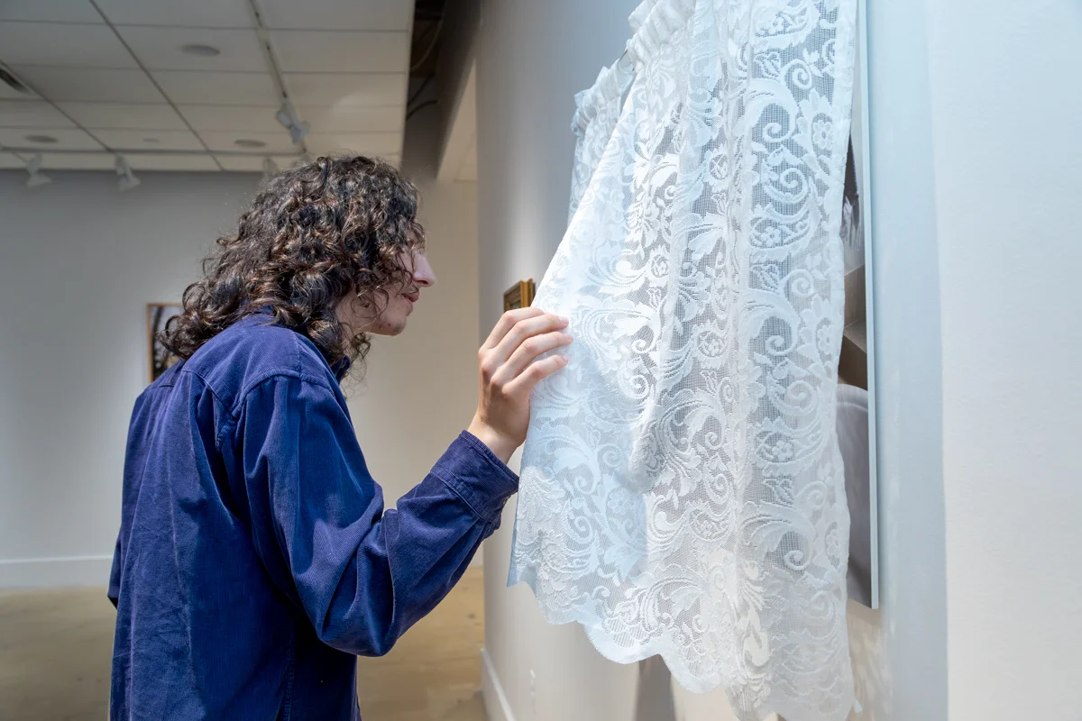 A person stands in a gallery space gently lifting a sheer lace curtain that partially covers a framed photographic artwork mounted on a white wall. The individual is shown in profile, wearing a long-sleeve blue shirt, with one hand raised to hold the lace fabric. The lace curtain features a floral pattern and obscures part of the image behind it. The gallery has white walls, polished flooring, and overhead track lighting, with additional artworks faintly visible in the background.