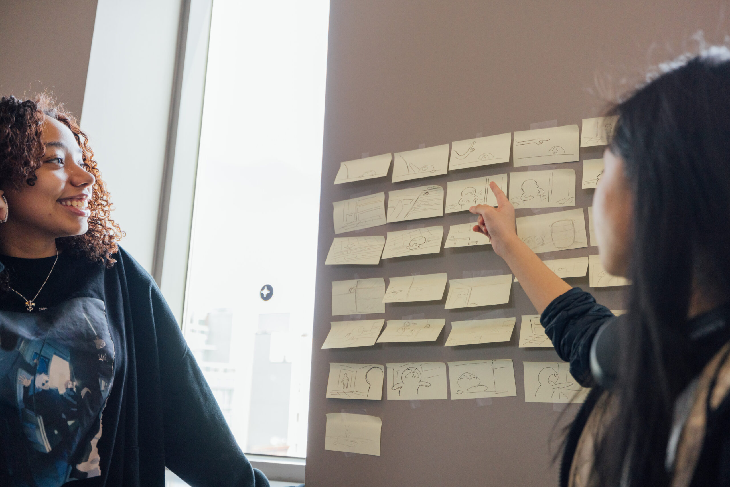 Two students stand beside a wall covered with rows of small sticky notes arranged in a grid, each containing hand-drawn storyboard sketches. One student points to a specific sketch while speaking, and the other smiles while looking at the board. The setting appears to be a classroom or studio space with natural light from a nearby window. The scene suggests a collaborative discussion or critique of storyboard concepts in a storytelling or animation course.