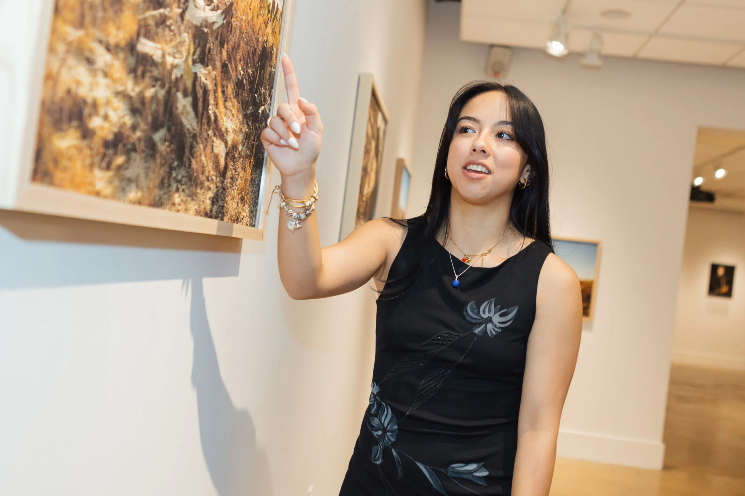 A person stands in a gallery space pointing toward a framed photographic artwork mounted on a white wall. The individual wears a sleeveless black top and jewelry on one wrist, with their arm raised to indicate a specific detail near the upper edge of the artwork. Additional framed photographs are visible along the wall behind them, and overhead gallery lighting illuminates the exhibition space.