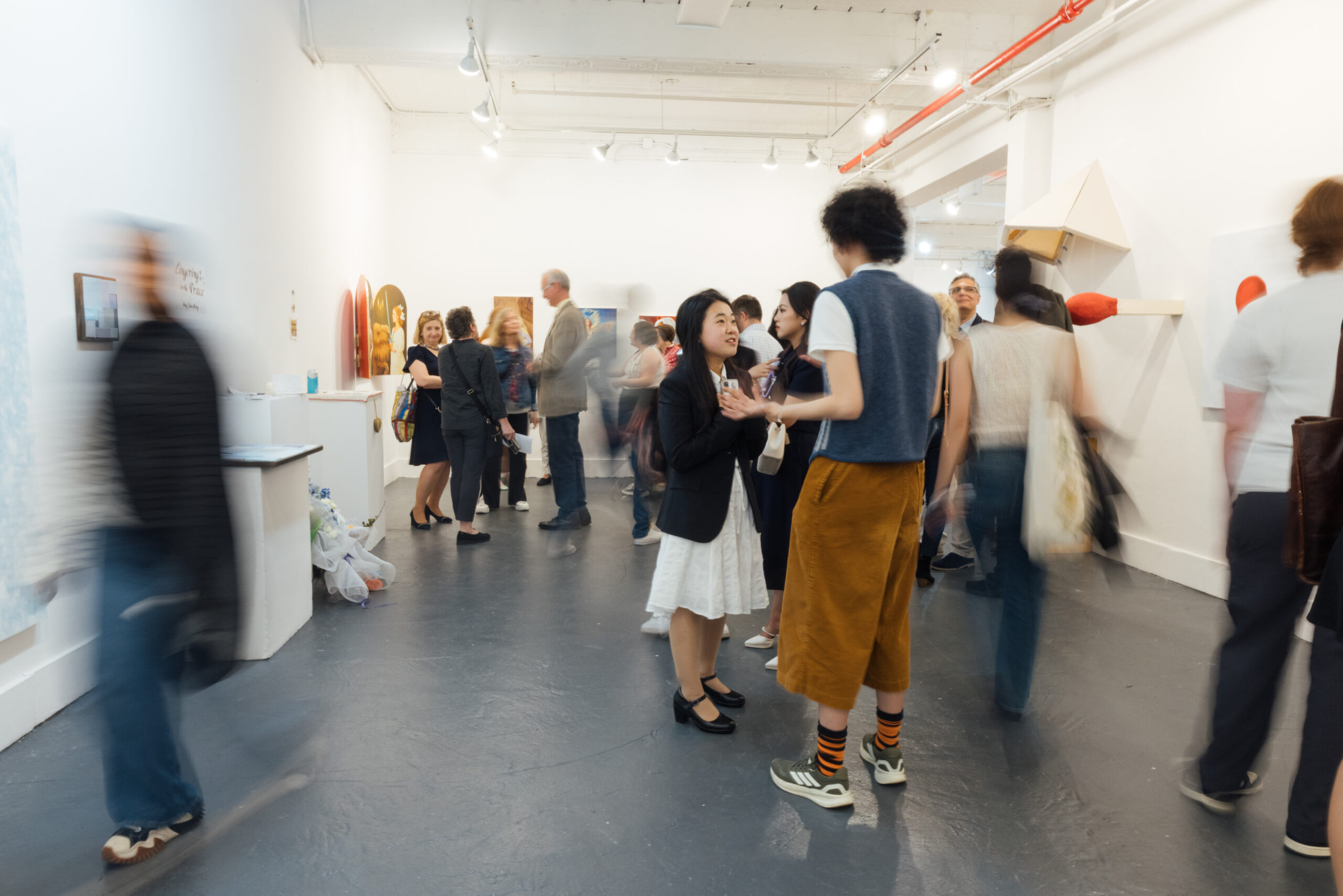 A crowded gallery scene during an art exhibition, with visitors standing, walking, and conversing throughout a white-walled exhibition space. Several figures appear in motion blur, suggesting movement and activity, while others stand still discussing artwork. Sculptural and wall-mounted artworks line the walls, including geometric forms and colorful pieces. Overhead track lighting illuminates the space, and exposed ceiling elements and red piping are visible above. The floor is dark gray, contrasting with the bright gallery walls.