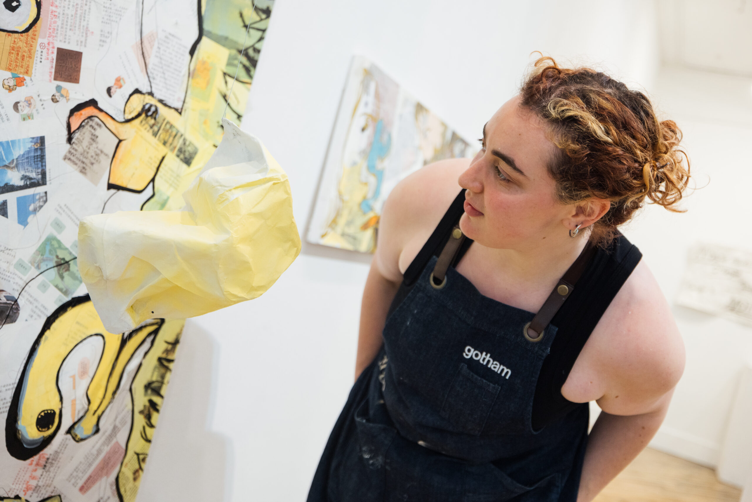 A person wearing a dark work apron leans in closely to examine a mixed-media artwork mounted on a white gallery wall. The artwork features layered collage elements, including printed text, drawings, and a protruding sculptural form made of translucent yellow material extending outward from the surface. Additional abstract artworks are visible blurred in the background, indicating a contemporary art exhibition setting.