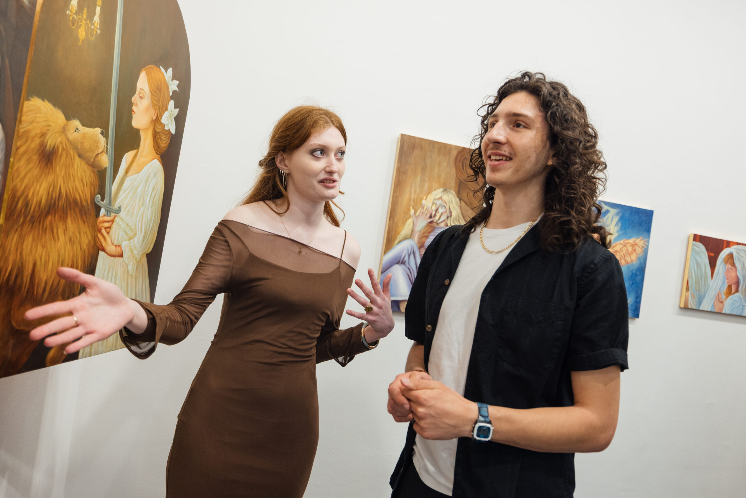 Two gallery visitors stand in conversation in front of a series of figurative paintings displayed on a white wall. One person gestures expressively with both hands while speaking, and the other listens attentively. The artworks behind them include painted portraits and symbolic imagery, including a large painting of a woman holding a sword beside a lion. The scene conveys active discussion and engagement within a contemporary art exhibition.