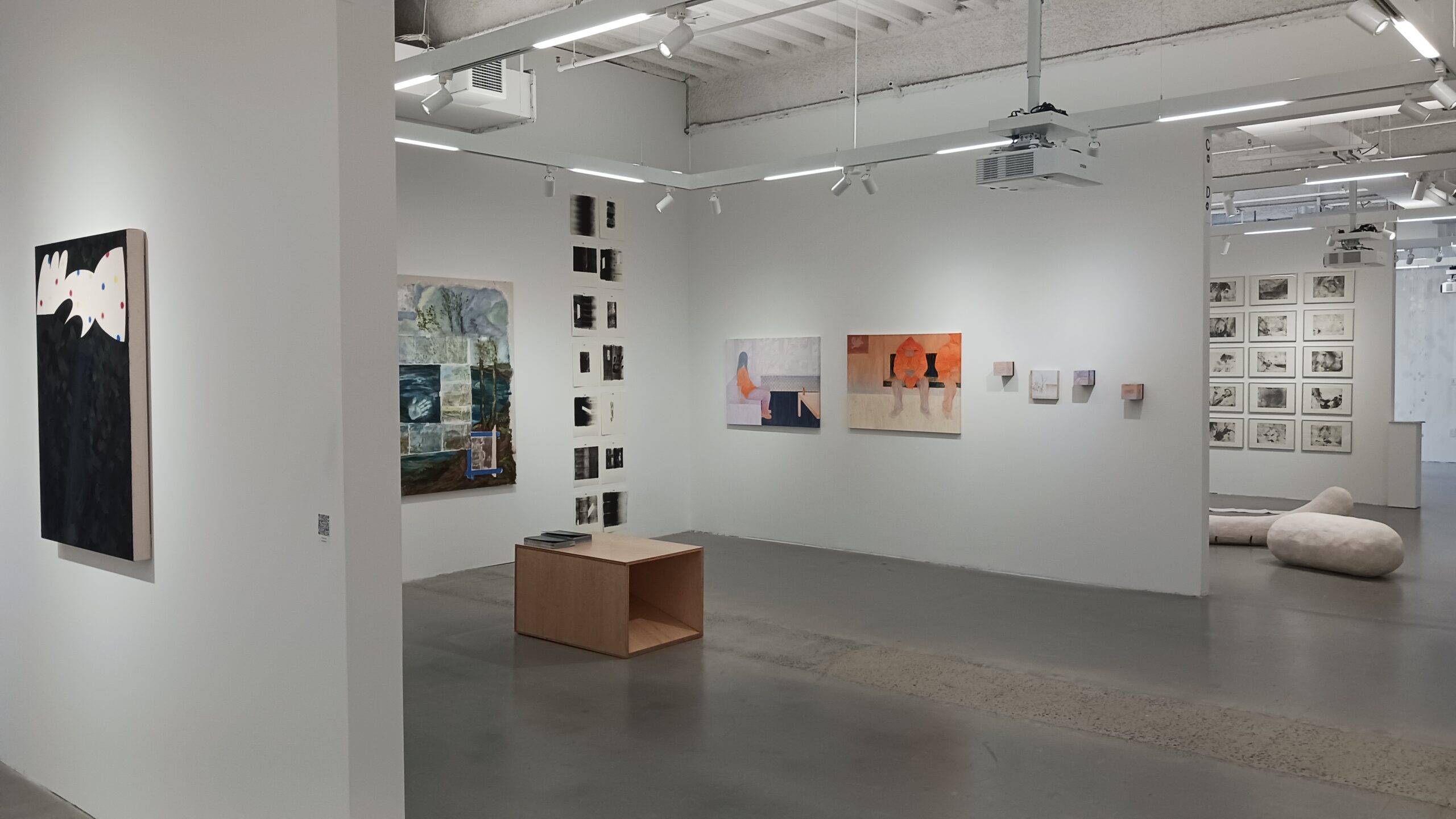An art gallery interior featuring several artworks on white walls. On the left, there is an abstract painting with black and white elements. Further along, a large collage-style artwork integrates various images. In the center, a light wood table sits on a concrete floor. To the right, two paintings depict figures, with one in orange tones, alongside smaller artworks. A series of framed images hangs on the wall in the background, and a soft sculpture is placed on the floor in the corner.