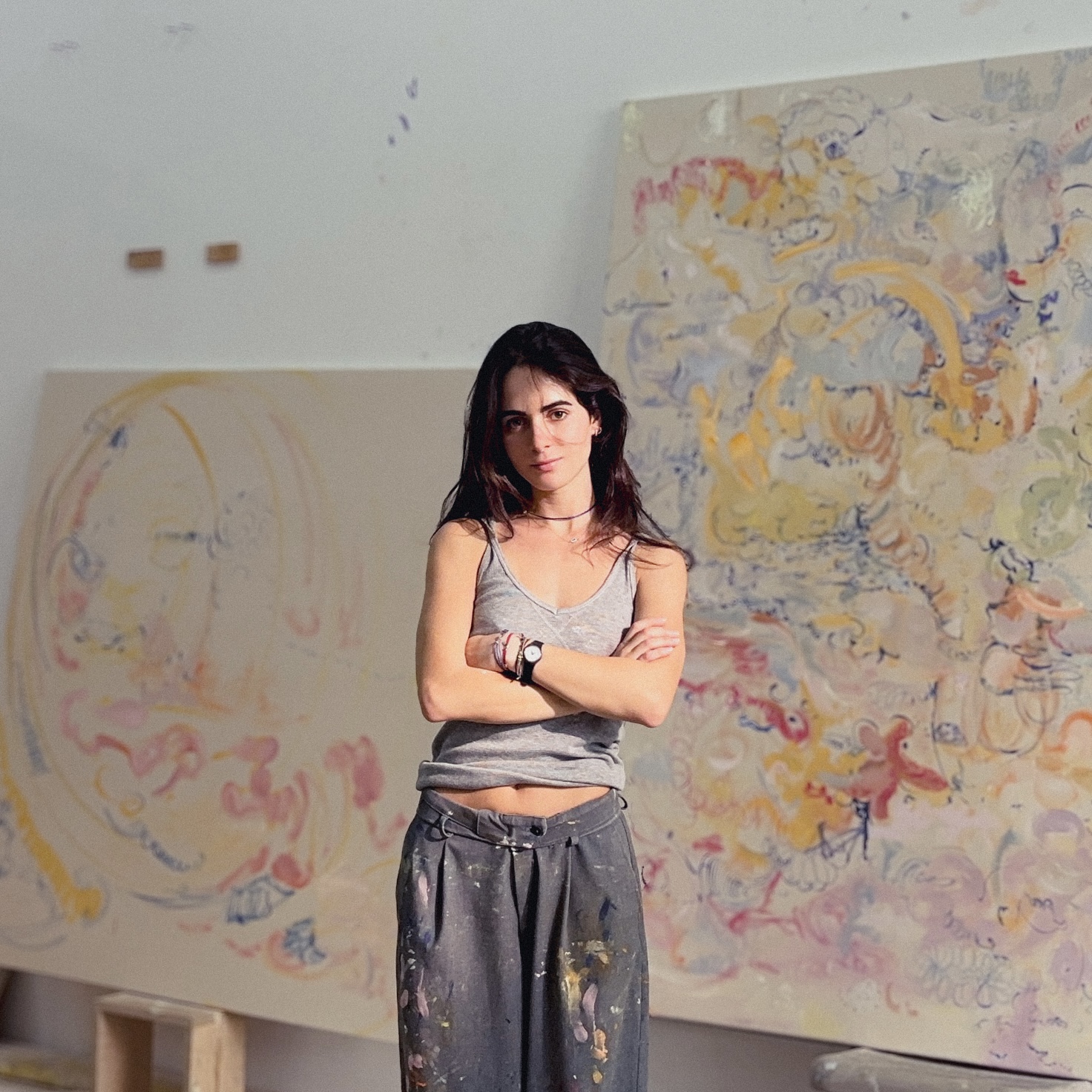 Artist Leda Tsoutreli stands with her arms crossed in front of two large canvases.