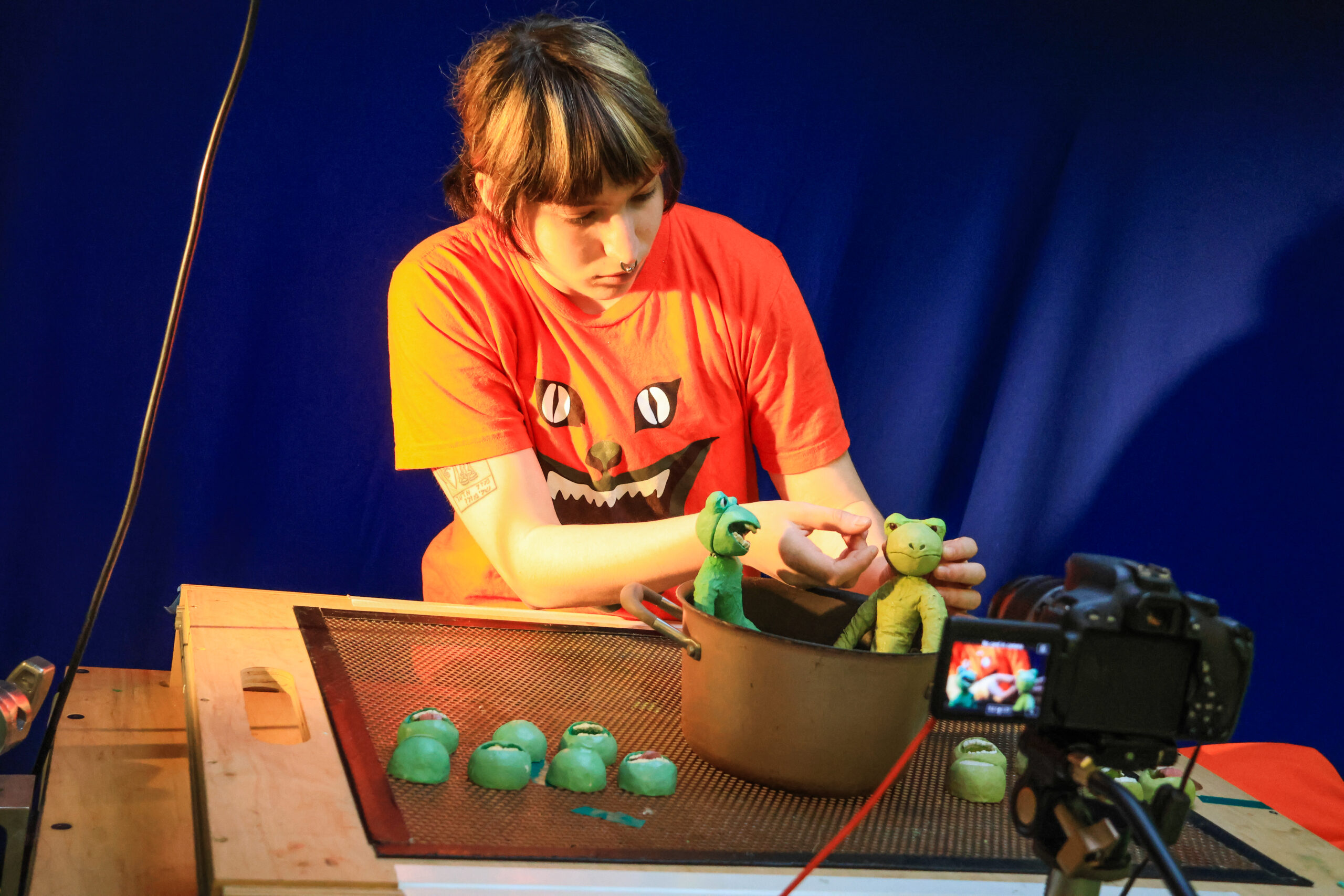 A person wearing an orange shirt with a cartoonish monster design is seen manipulating green clay figures while seated at a wooden table. In front, there are several green clay heads and a brown pot containing two larger clay characters. A camera on a tripod is focused on the scene. The background is a blue fabric, and the lighting highlights the workspace's details.