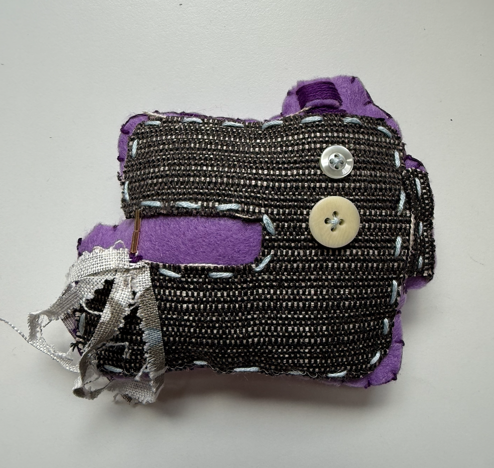 A small, handmade plush item resembling a camera, made from various fabrics including dark woven material and purple felt. It features decorative stitching, two buttons, and a small metal clip. The edges are slightly frayed, giving it a textured appearance.