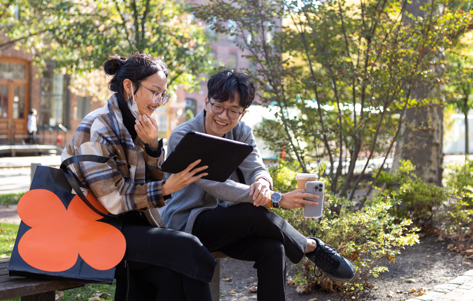 Two students sit on a bench outside on campus. One is holding a tablet which they are both looking at. The other is holding a coffee.